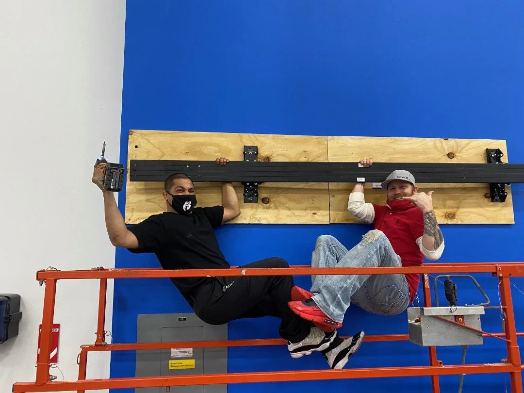 Two men sitting on a scissor lift in front of a wall with a blue background. One man is holding a drill, wearing black clothes and a face mask, while the other man is wearing a red shirt, a cap, and is smiling.