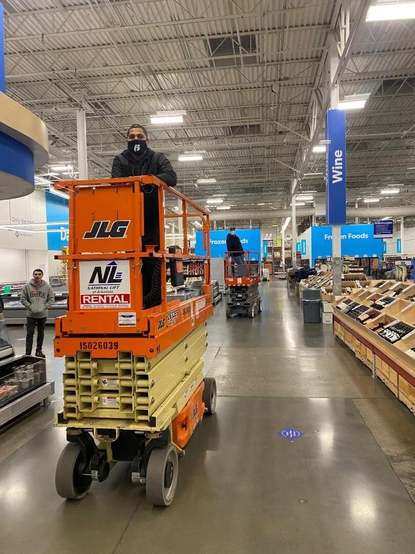 Worker standing on an orange scissor lift inside a grocery store aisle with wine and frozen food sections visible.