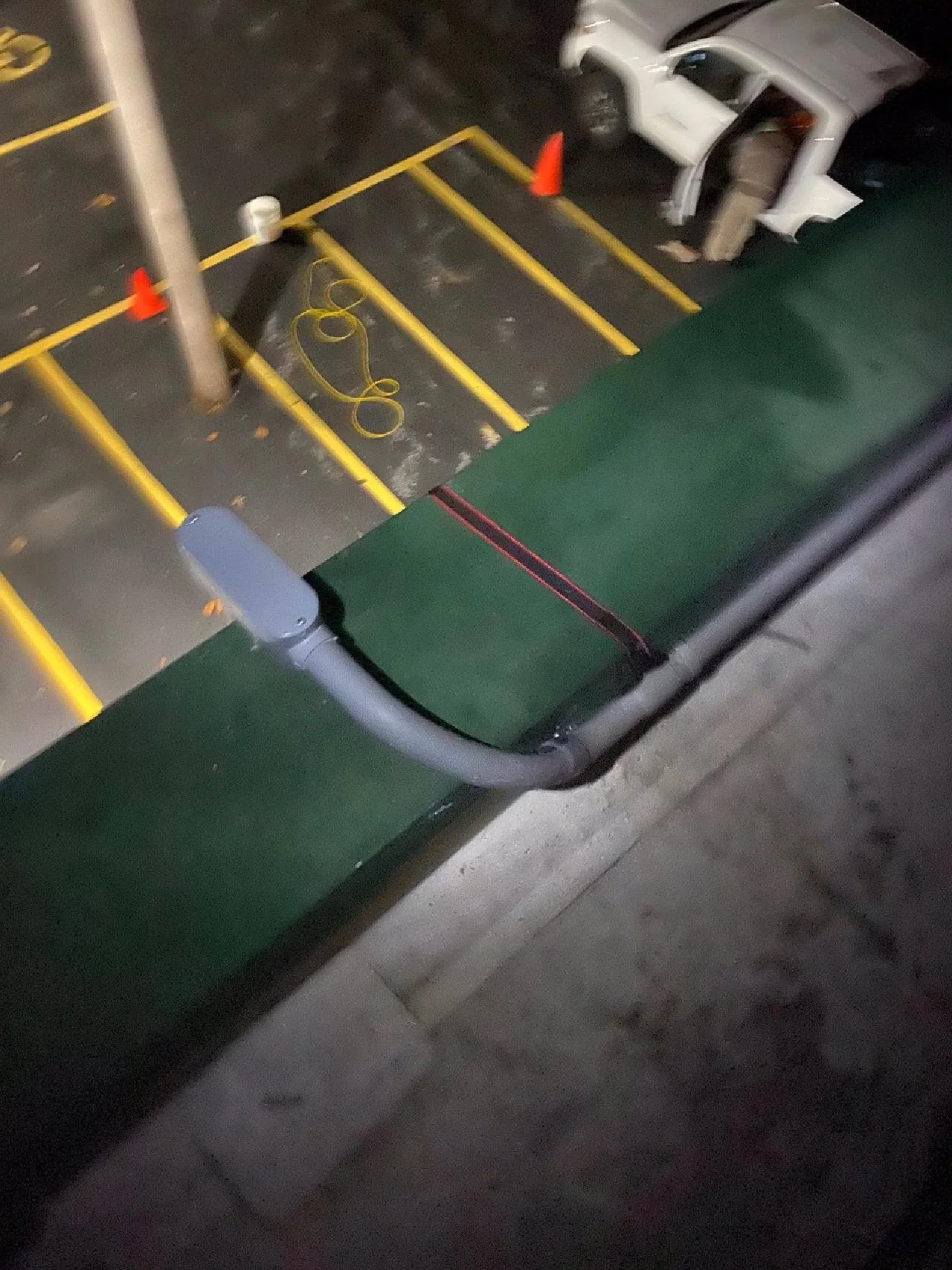 Nighttime parking lot with an overturned white truck, yellow parking lines, orange cones, a cord, and a green structure at the edge.