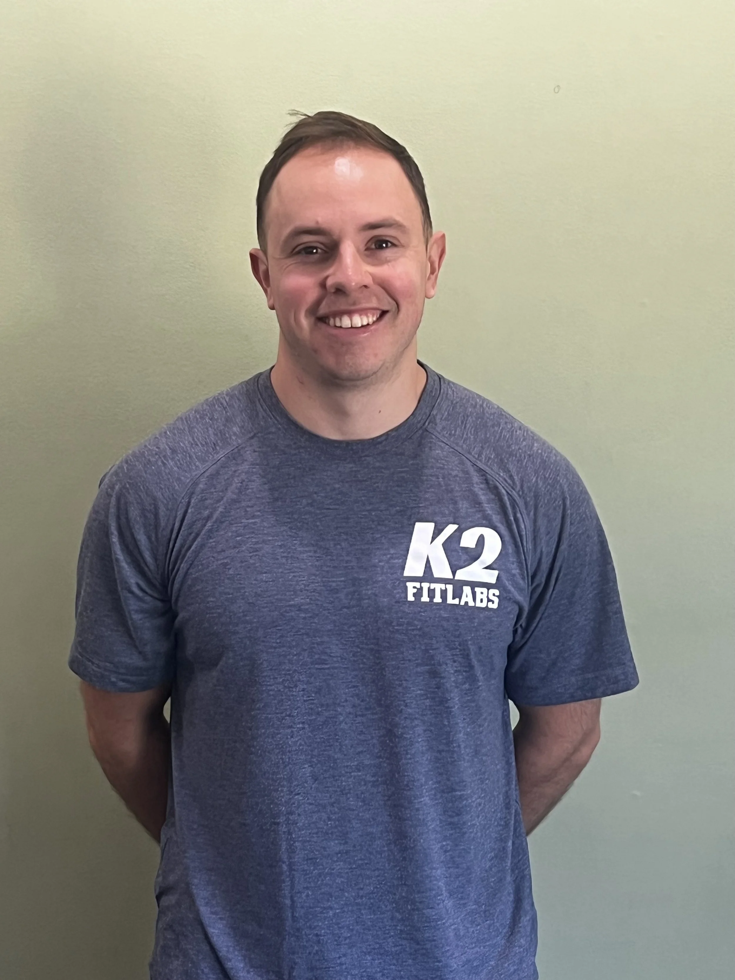 A smiling man wearing a gray athletic T-shirt with "K2 FITLABS" logo stands against a plain light green wall.