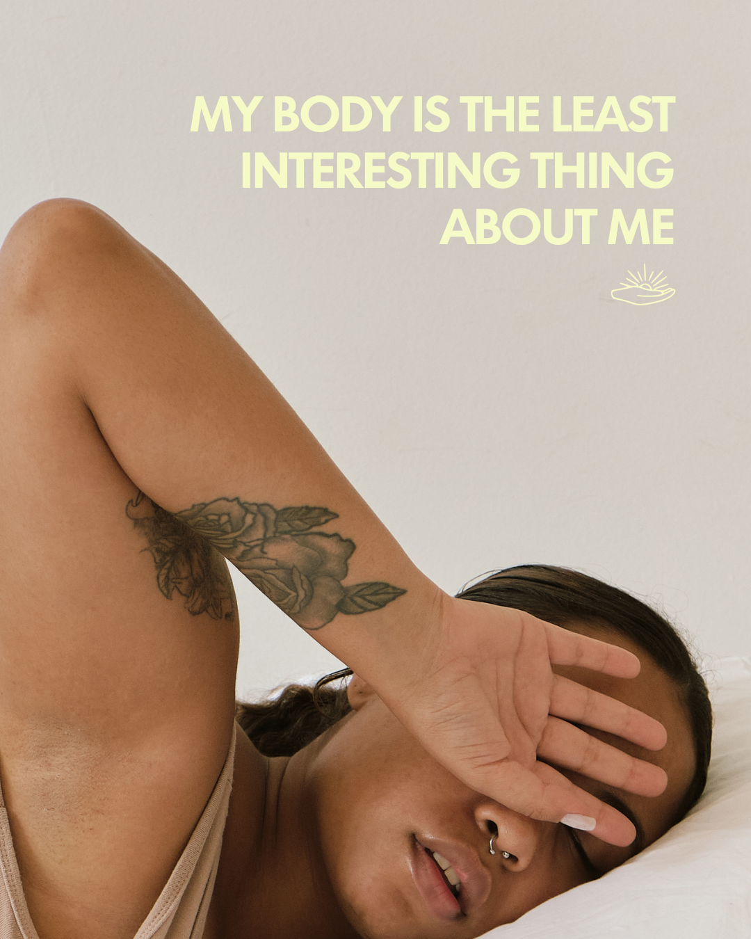 A person lying on a bed with their hand covering their face, featuring a tattooed arm. The person has a septum piercing and is smiling slightly. There is a quote in pale yellow text at the top that says, "My body is the least interesting thing about 