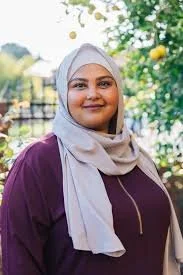 Woman wearing a white hijab and maroon top outdoors, smiling with trees and yellow flowers in the background.