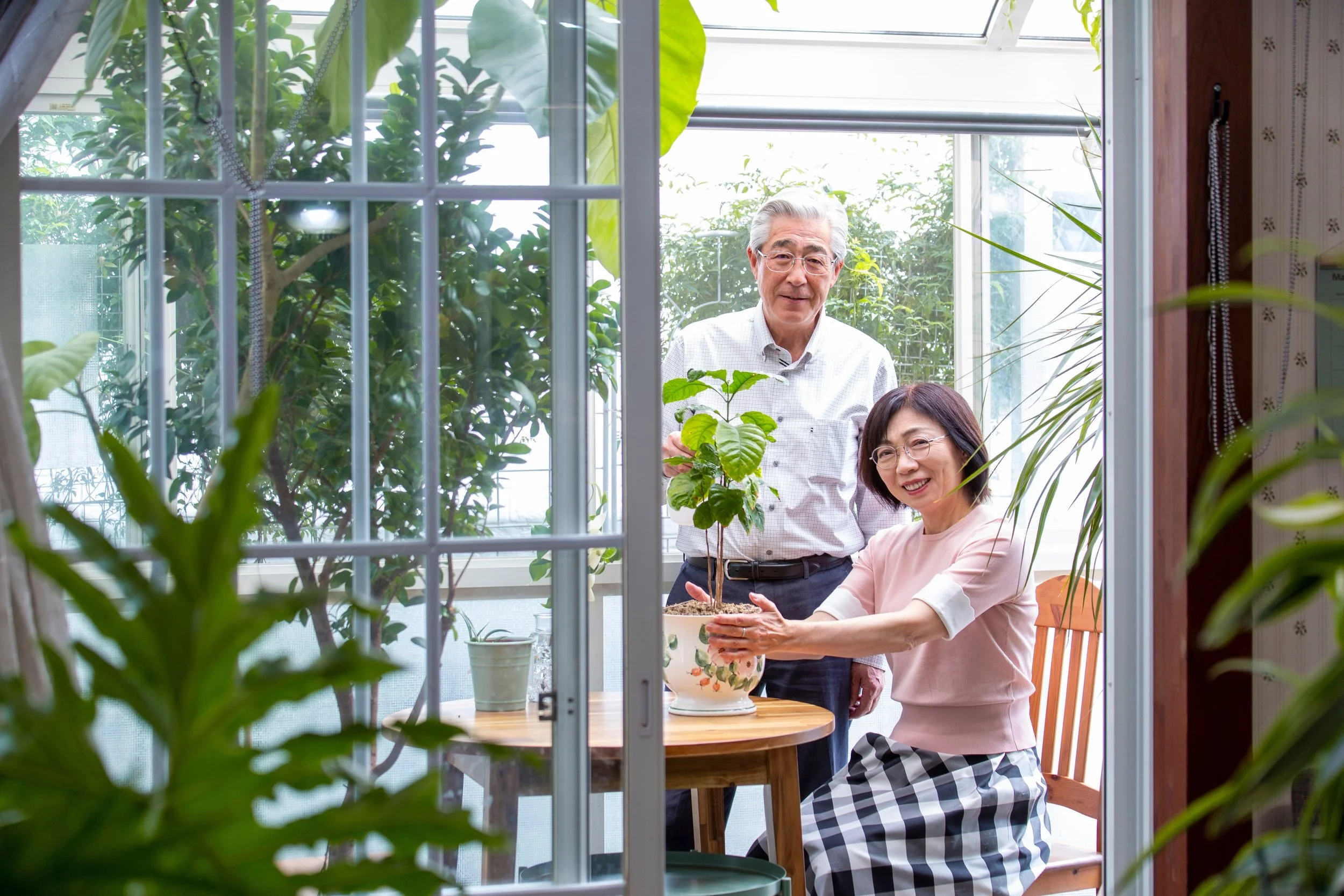 An older couple looking happy and relaxed, tending a plant together in a sunlit conservatory — representing coordinated planning through retirement.