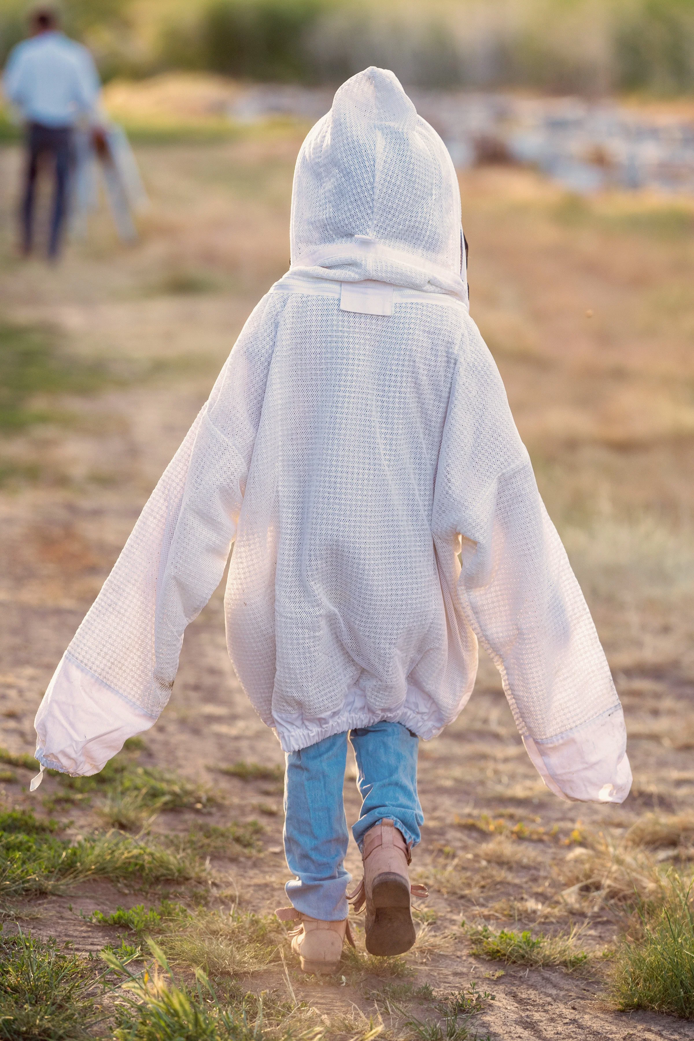 Child walking on a dirt path in a rural outdoor setting, dressed in a large white hoodie and blue jeans, with person in the background.