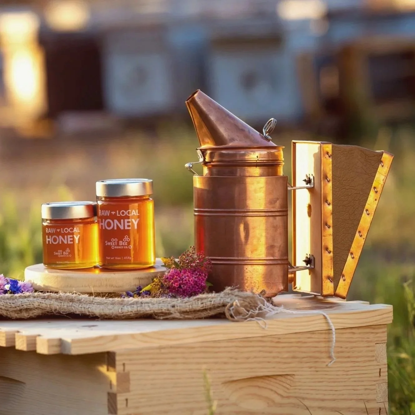 Honey jars labeled 'RAW + LOCAL HONEY' placed on a wooden surface with flowers, alongside a copper honey extractor in a rural outdoor setting.