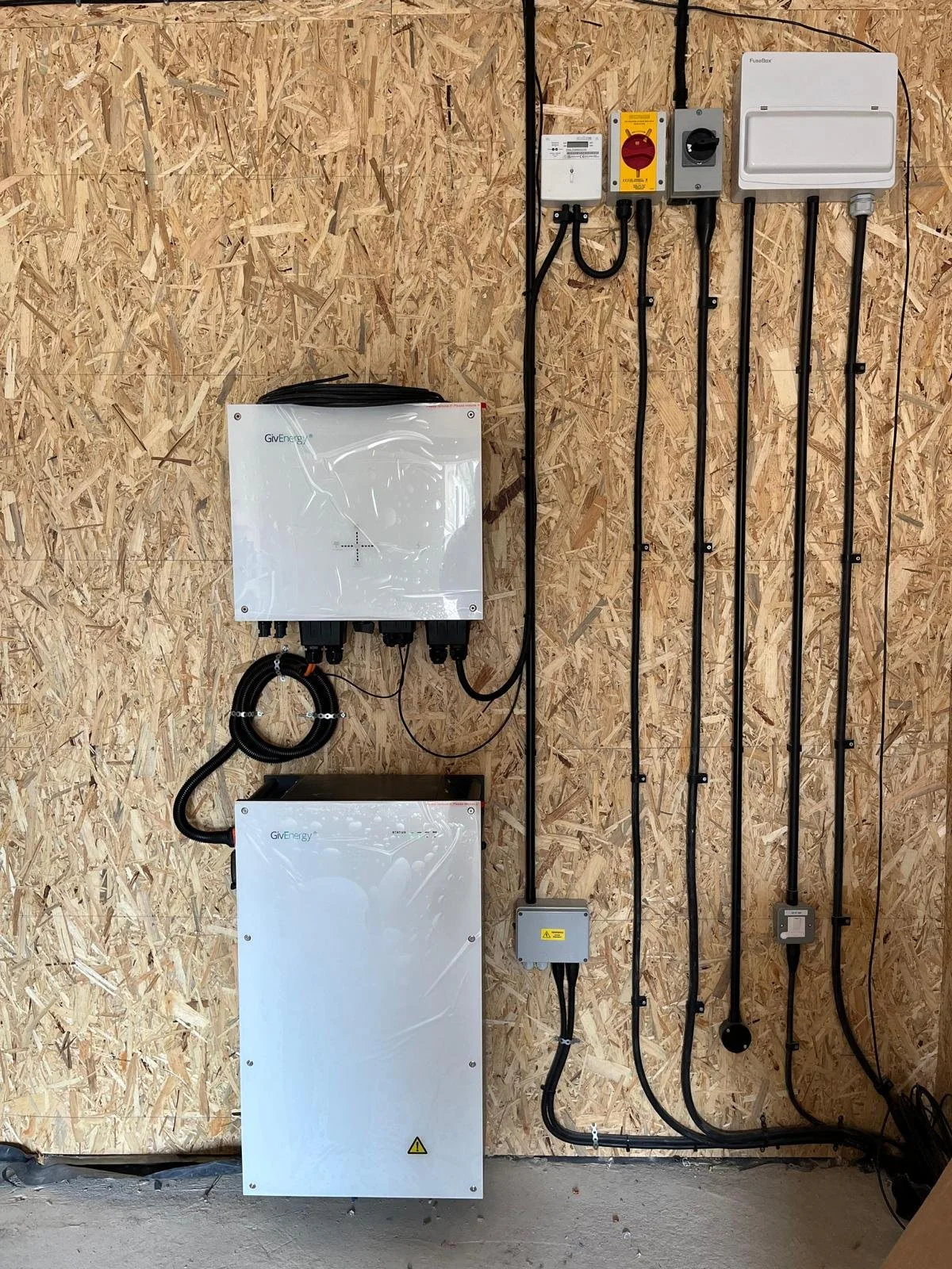 Electrical equipment mounted on a plywood wall, including solar inverter, disconnect switches, circuit breakers, and wiring for a solar power system.