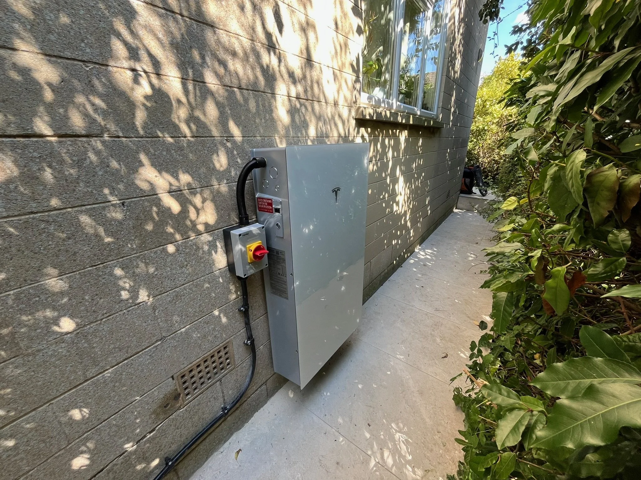 Exterior of a house with a window, alongside an electrical box on a beige brick wall, partly shaded by tree shadows and surrounded by green bushes.