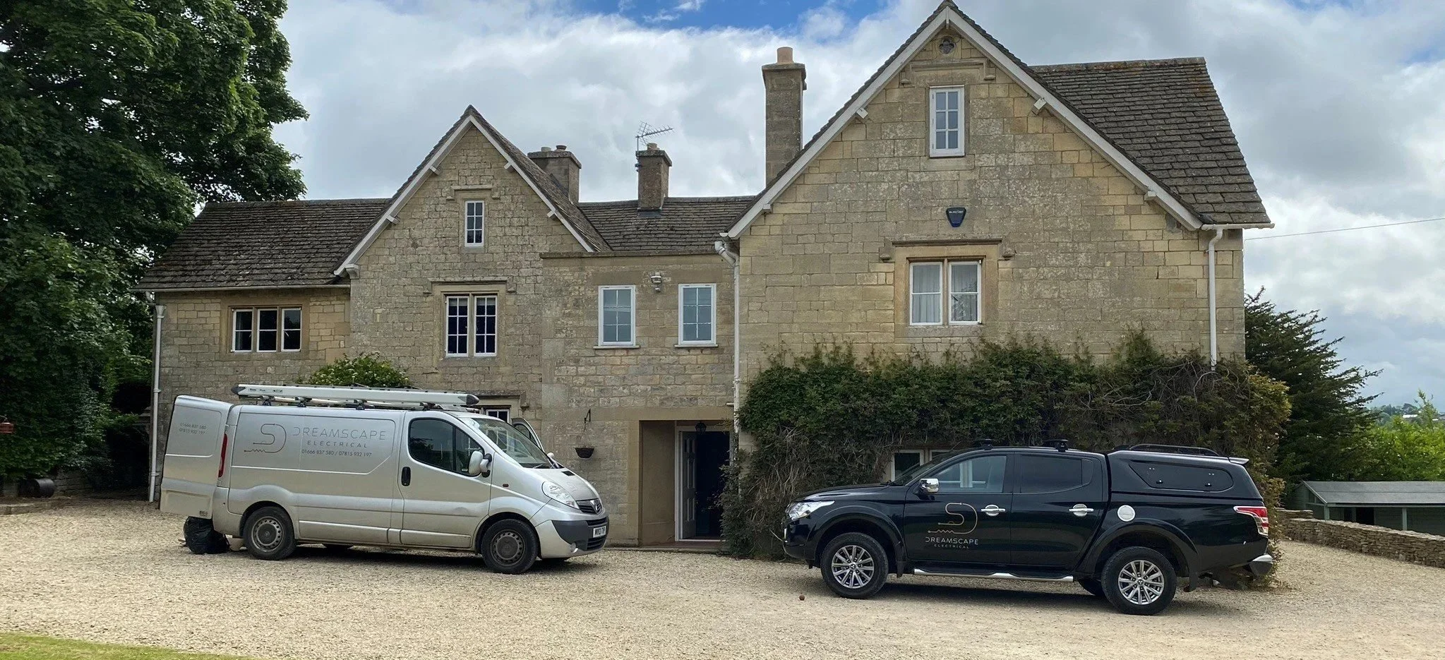 Stone house with multiple windows and a steep gabled roof, parked cars in front, including a white van and a black SUV, greenery around the house, cloudy sky above.