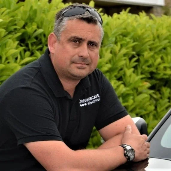 A man with short dark hair and a serious expression, wearing sunglasses on his head and a black polo shirt with the text 'Dreamscape Home Electrical,' sitting outdoors in front of green foliage.