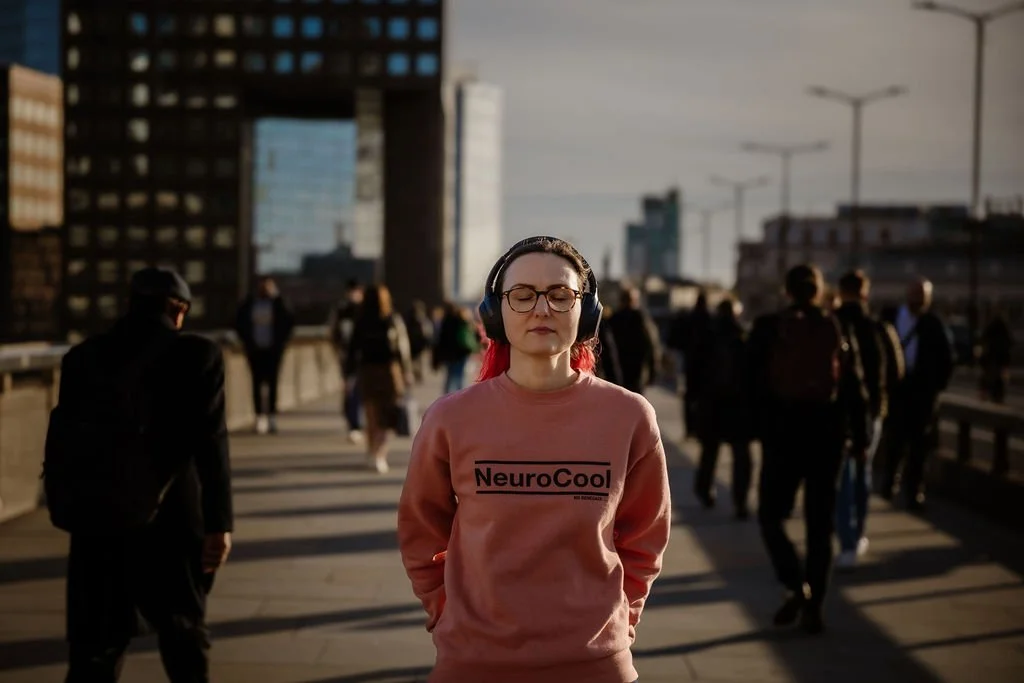 Founder of Expand the Circle, Alicja Nocon, standing in the middle of a busy walkway, wearing a pink jumper that says “Neurocool,” eyes closed and headphones on