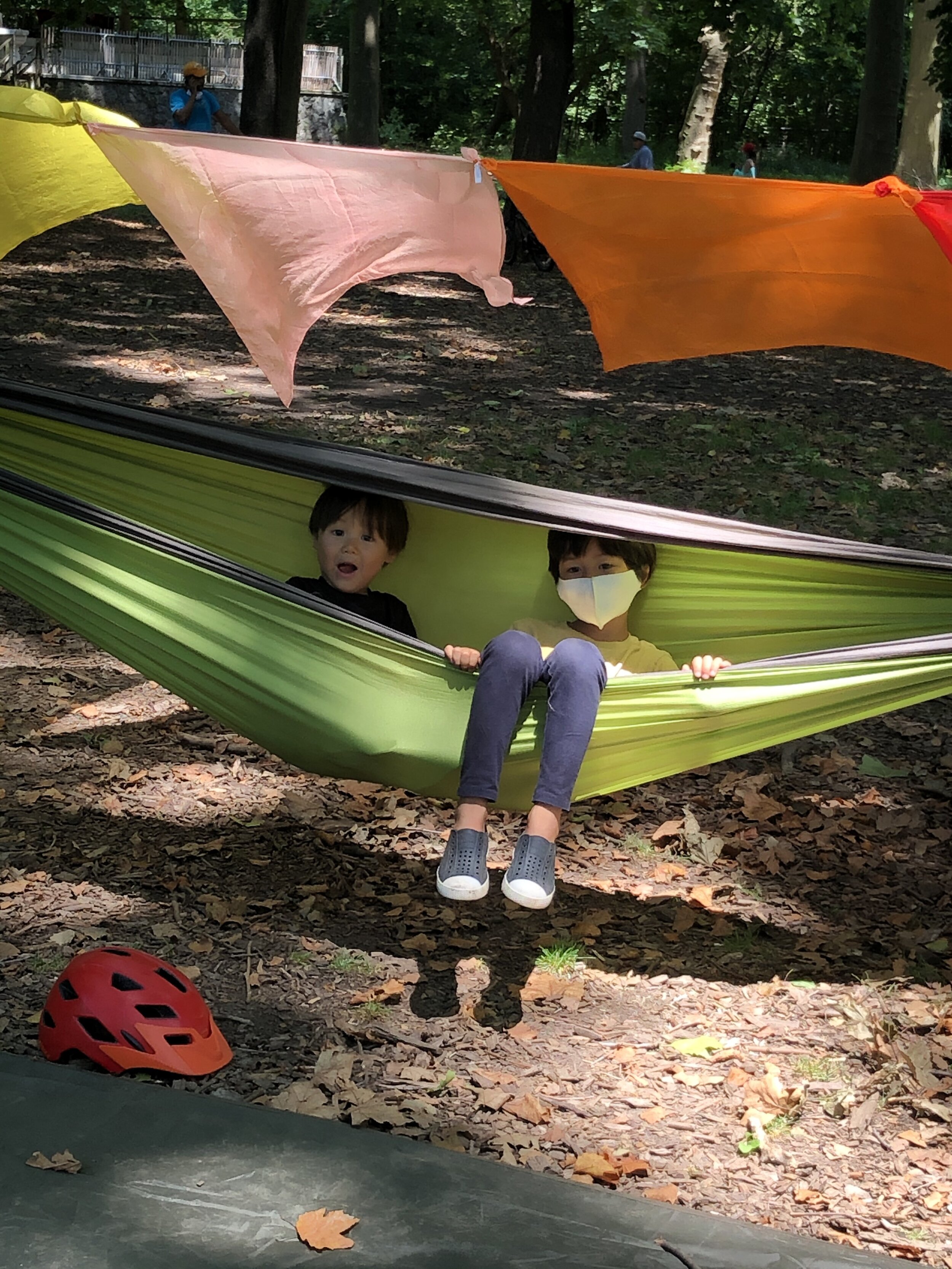 A close up photo of Honeytree Forest child Tree and his big brother Pau smiling while tuck into a green hammock in the shade of the Nethermead trees like peas in a pod. In the background, wave a line of large silk fabric squares tied together, each i