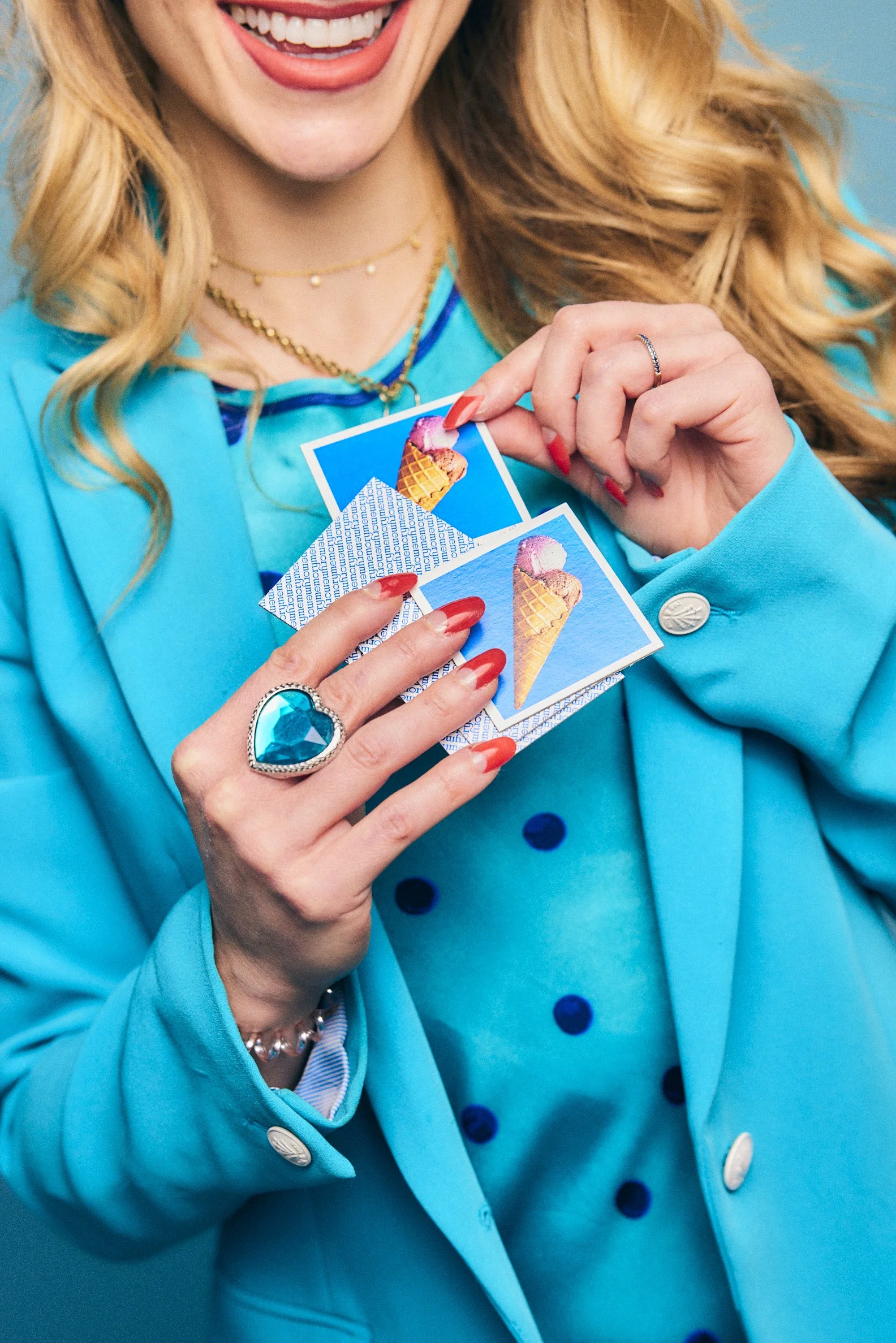 Close-up of a woman in a bright blue coat holding two photos of ice cream cones, smiling with red lipstick and wearing gold jewelry.