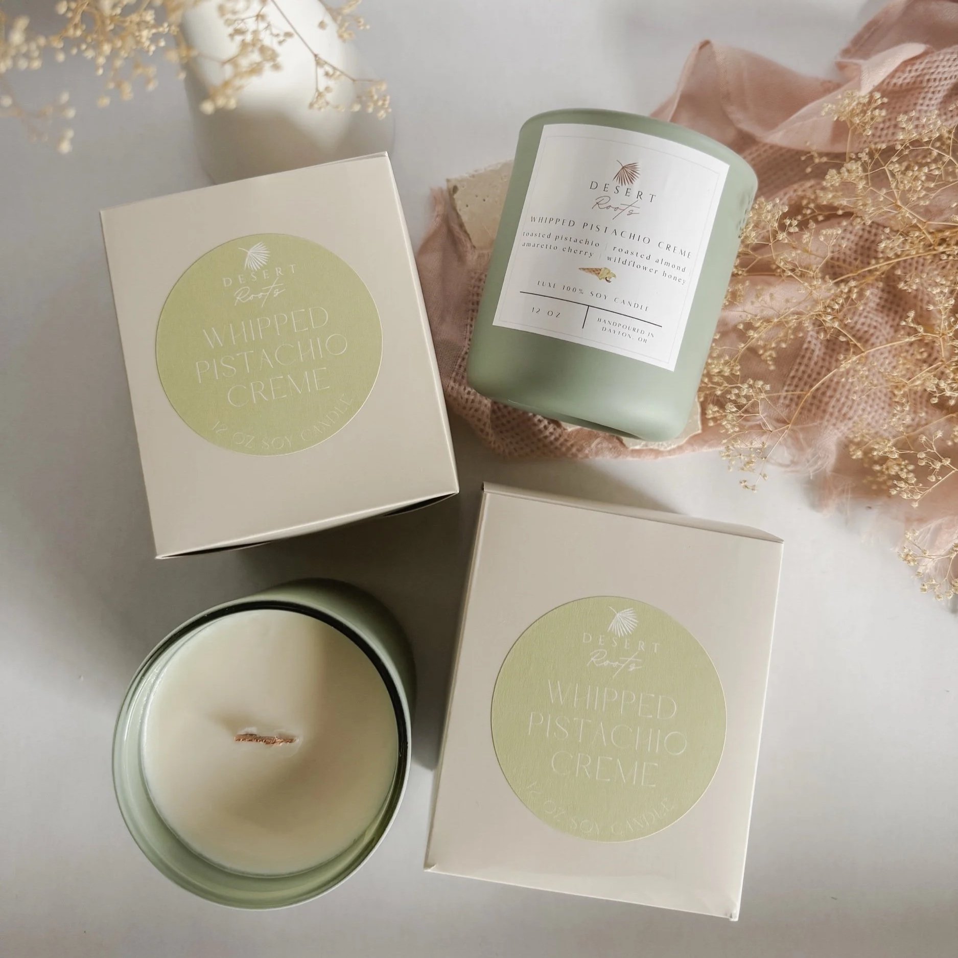 A white box and a pale green candle jar with labels that read 'Desert Roots' and 'Whipped Pistachio Creme,' placed on a soft pink lace cloth with dried flower sprigs.
