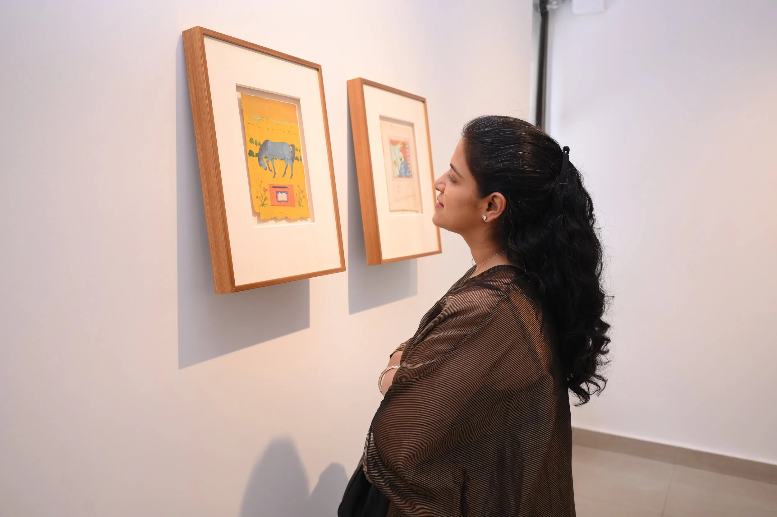 Woman with long dark hair, wearing a brown jacket, viewing artwork in a gallery.