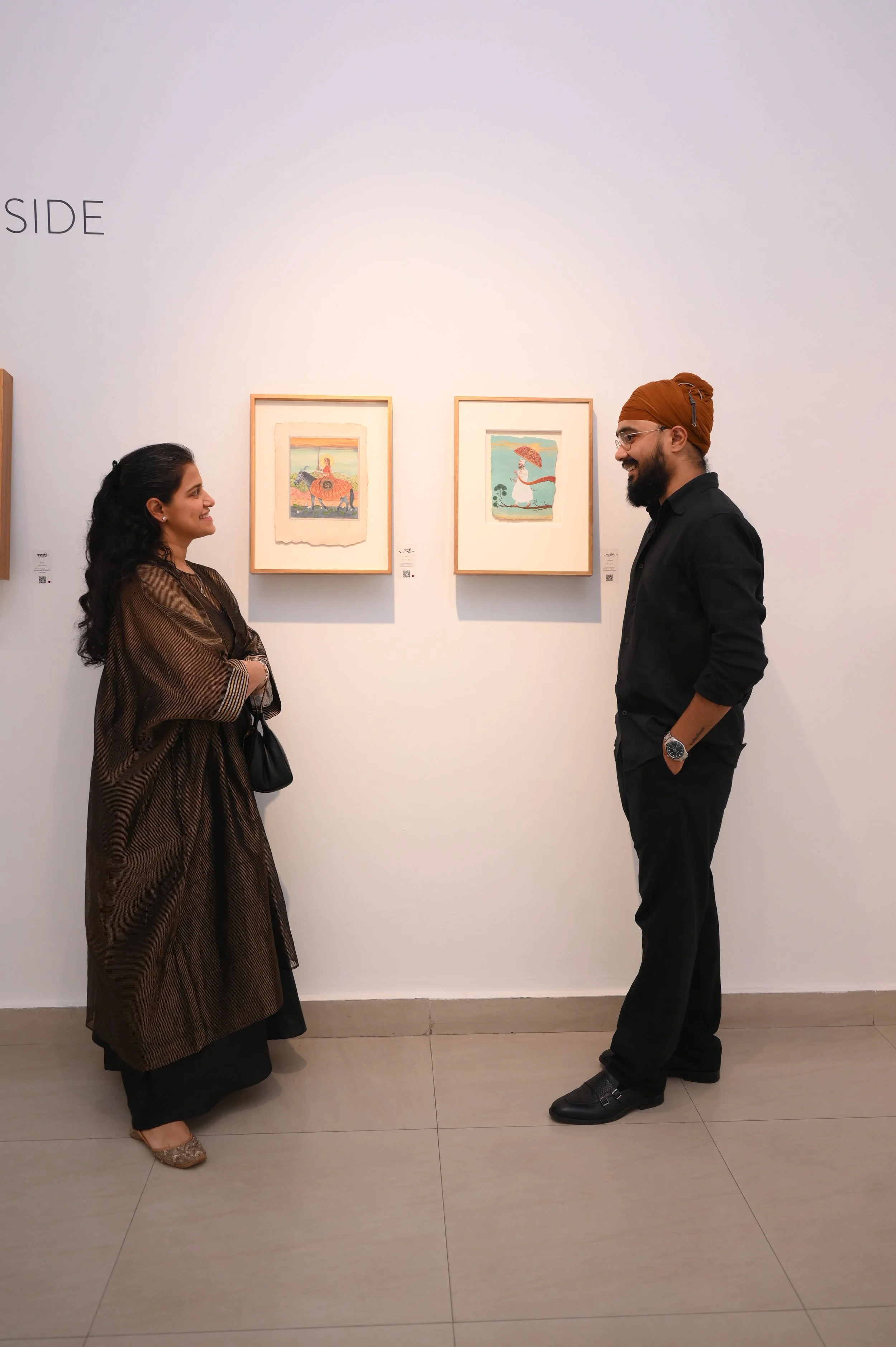 Ravjot singh standing and looking at the woman in an art gallery. The woman is wearing a long, sheer brown dress with striped sleeves and the man is dressed in black with a turban and glasses. There are two framed artworks on the wall behind them.