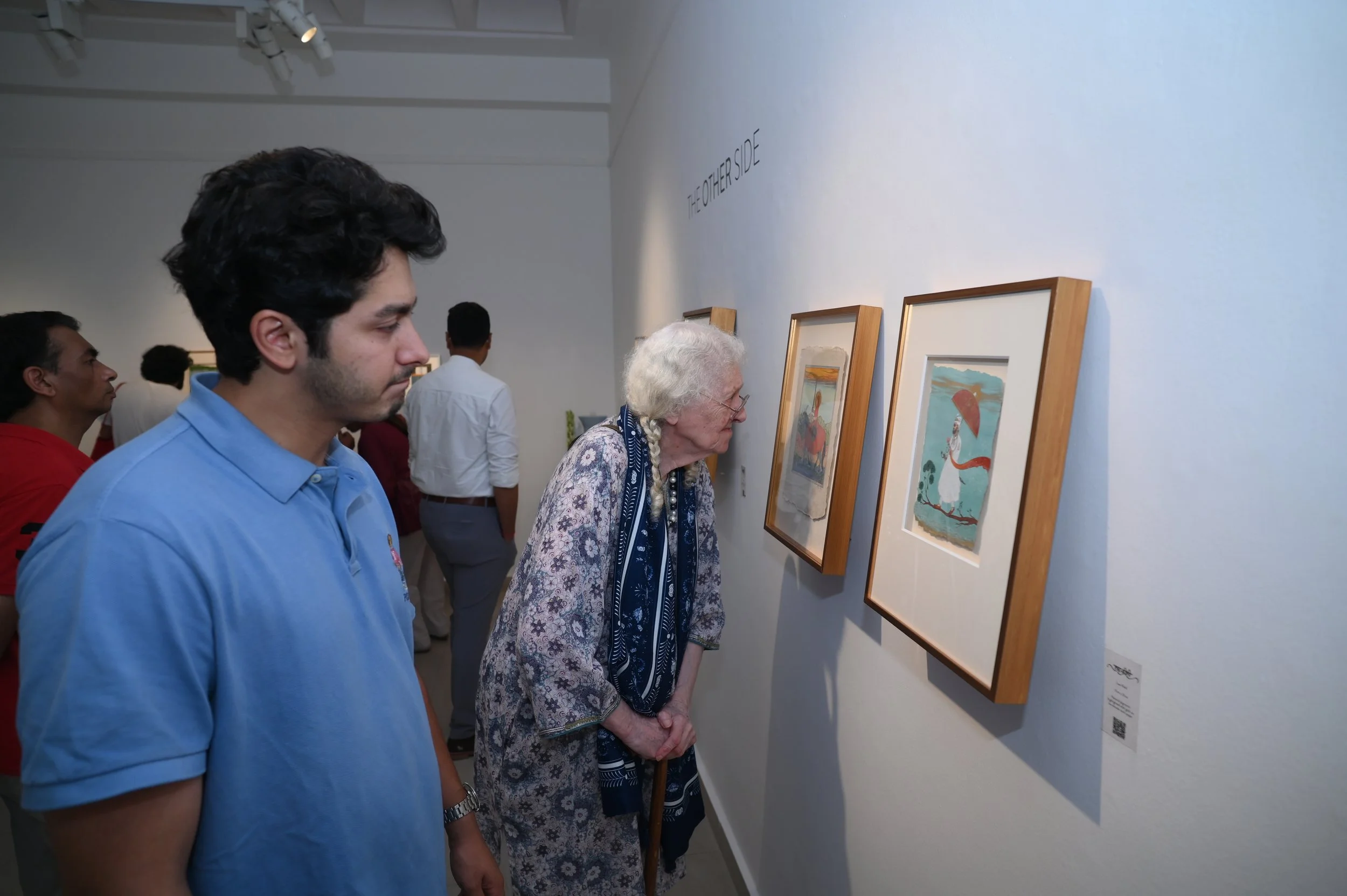 Multiple people viewing framed artwork in an art gallery, including an older woman with a cane and a younger man in a blue shirt.