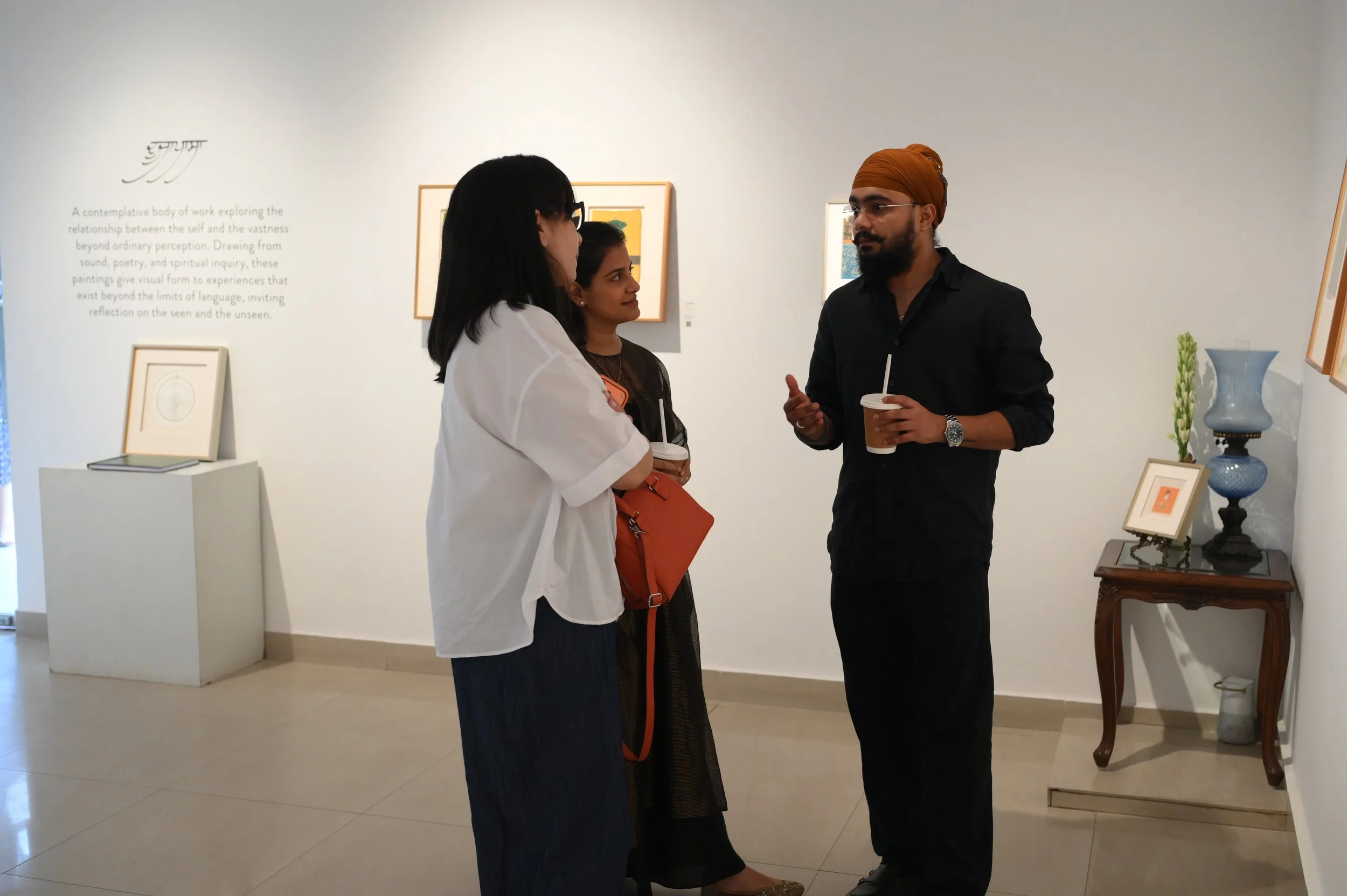 Ravjot singh having a conversation with two women, one wearing glasses and the other with long dark hair, and a man with a turban and glasses, are holding drinks. The background features framed artworks on the wall and a table with decorative items.