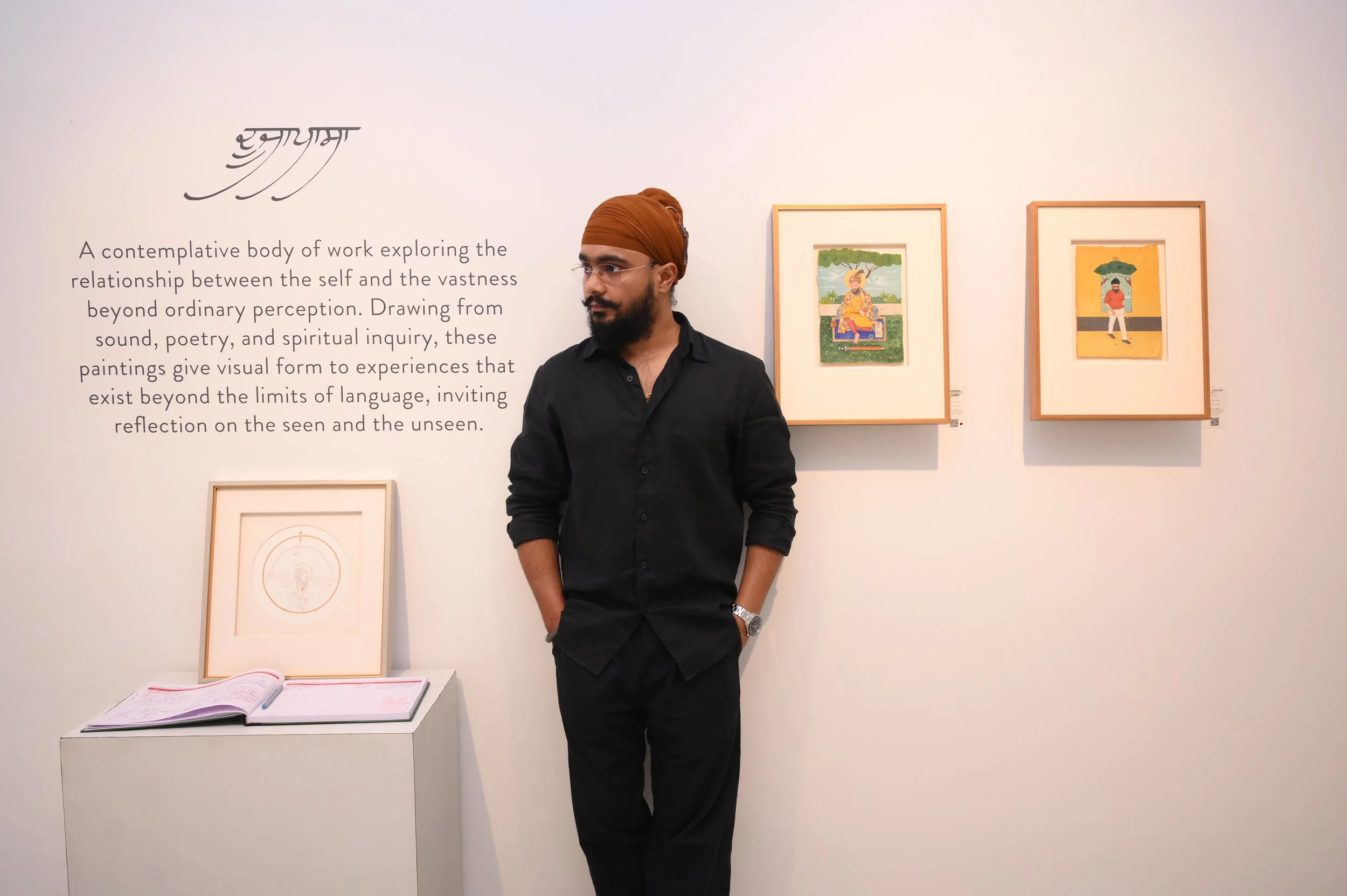 Ravjot Singh stands with his hands in his pockets in an art gallery. Behind him, there are framed paintings on the wall, and an open book lies on a white pedestal.