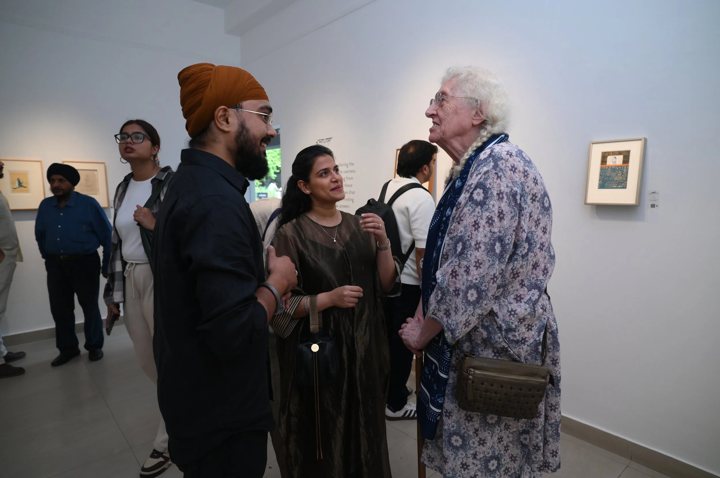 A diverse group of five people gathered in an art gallery, engaging in conversation. The background features framed artwork on the white walls.
