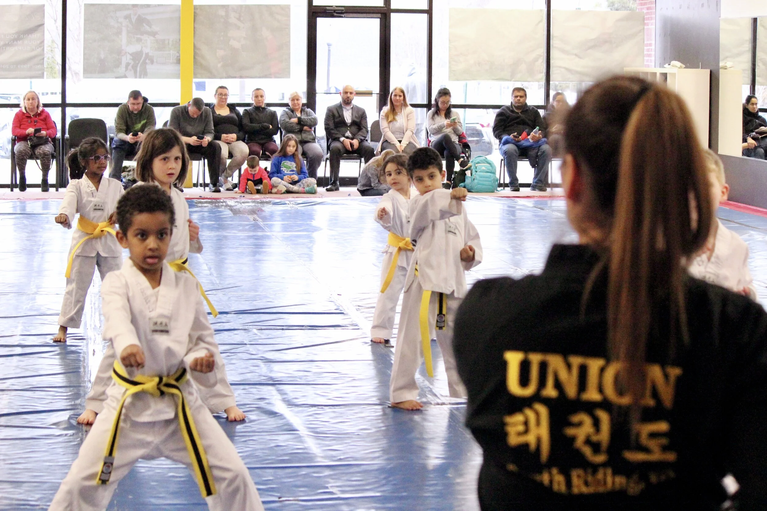 Children in martial arts uniforms practicing kicks on a blue mat while spectators sit in chairs behind a large glass storefront window, and an instructor in a black uniform with yellow lettering stands in the foreground.