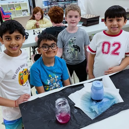 Four children smiling at a science activity table with a jar and a container of colored liquid, with two additional children in the background at a different table.