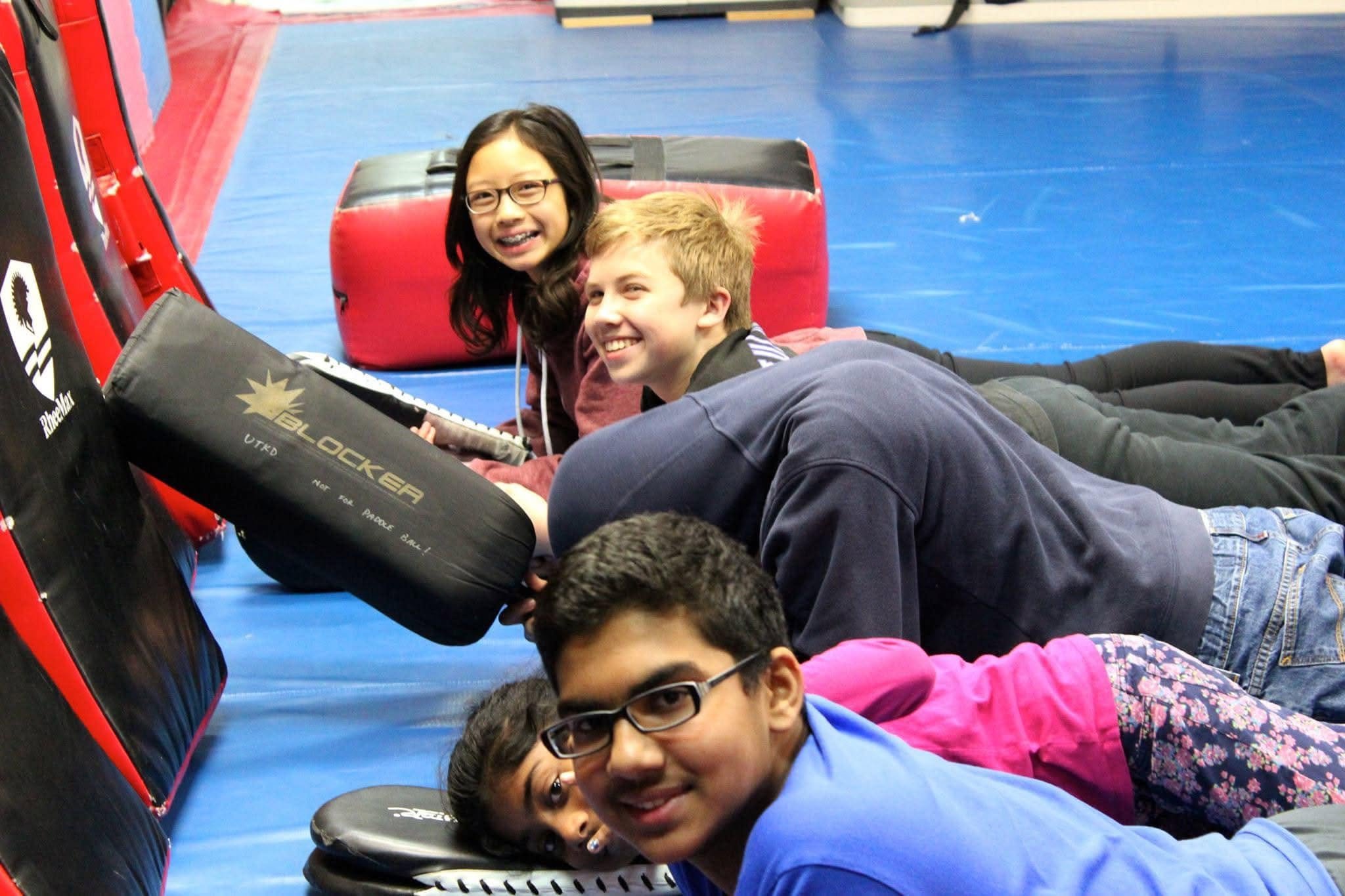 Group of children lying on the floor in a gym, smiling and playing around a boxing punching bag.