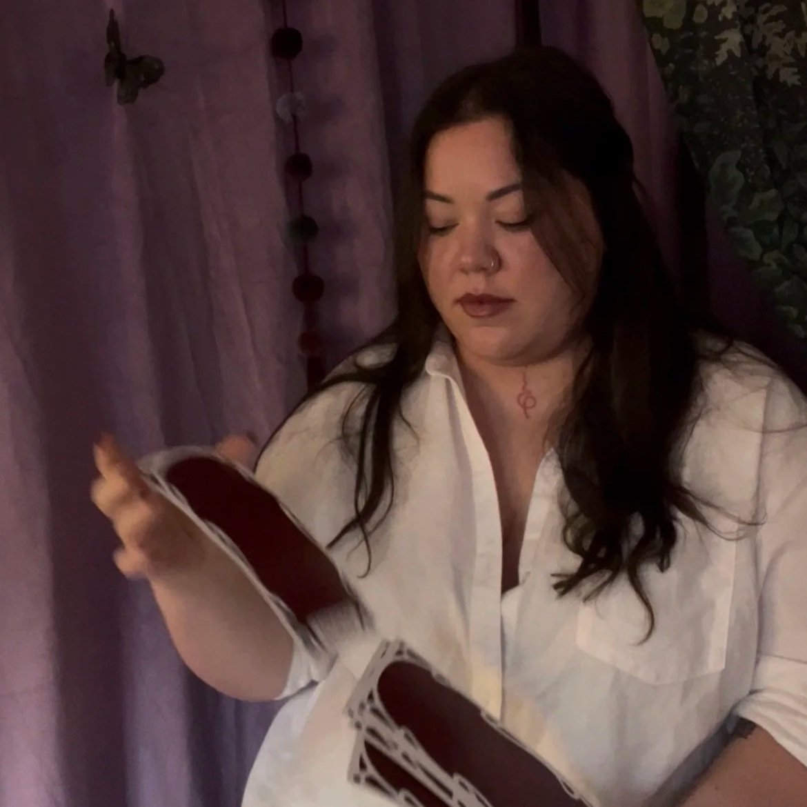 A young woman with dark hair and a nose piercing reading a fan of tarot cards in a dimly lit room with purple curtains and string lights.