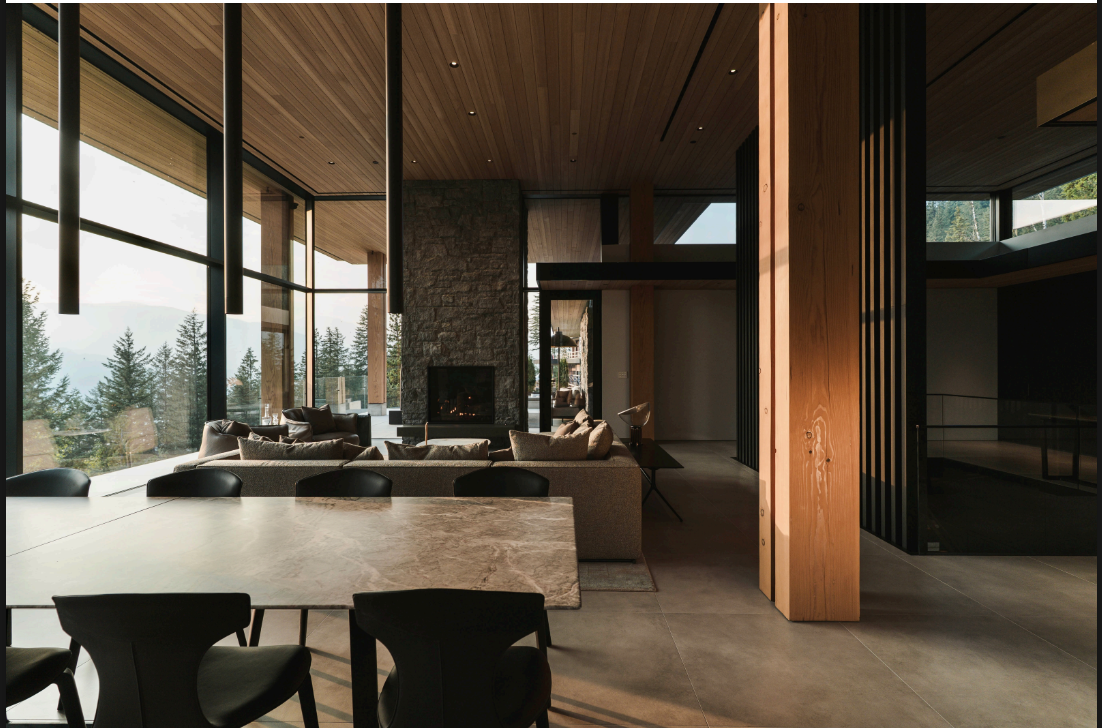Modern living room with large windows showing outdoor trees. Contains a stone fireplace, beige sofa, and a wooden ceiling.