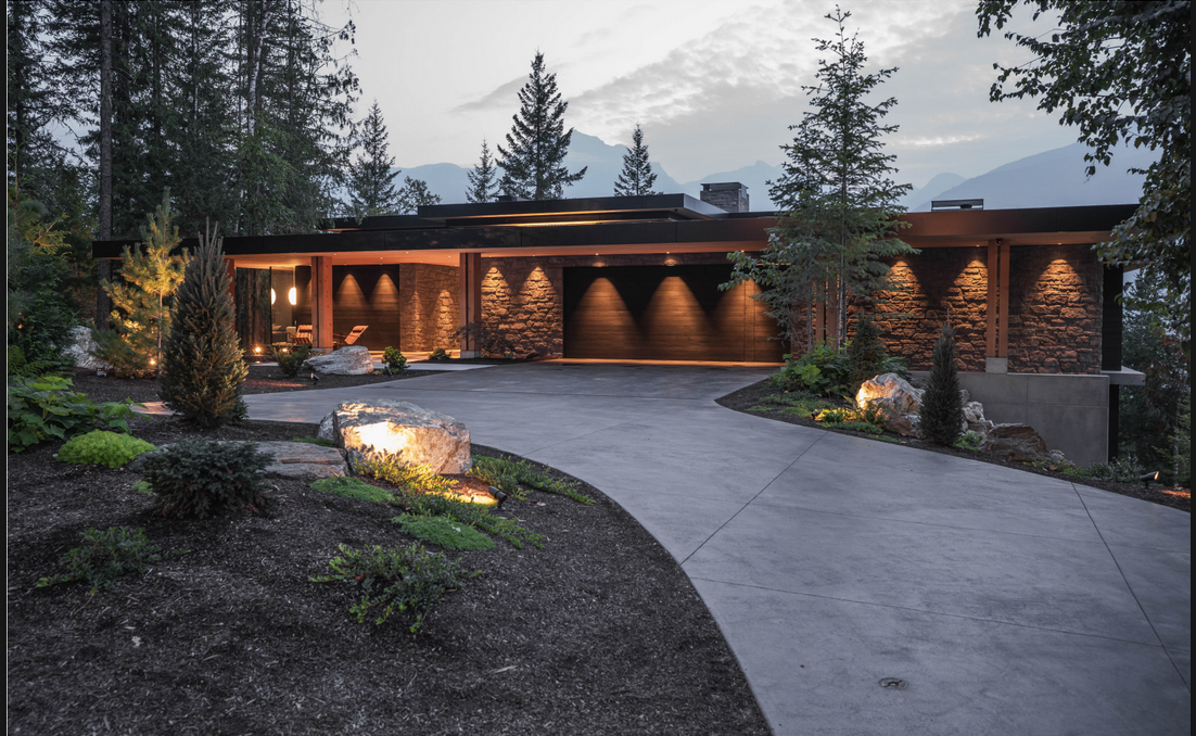 Modern house in a forested area with exterior lighting, trees, rocks, and a curved concrete driveway.