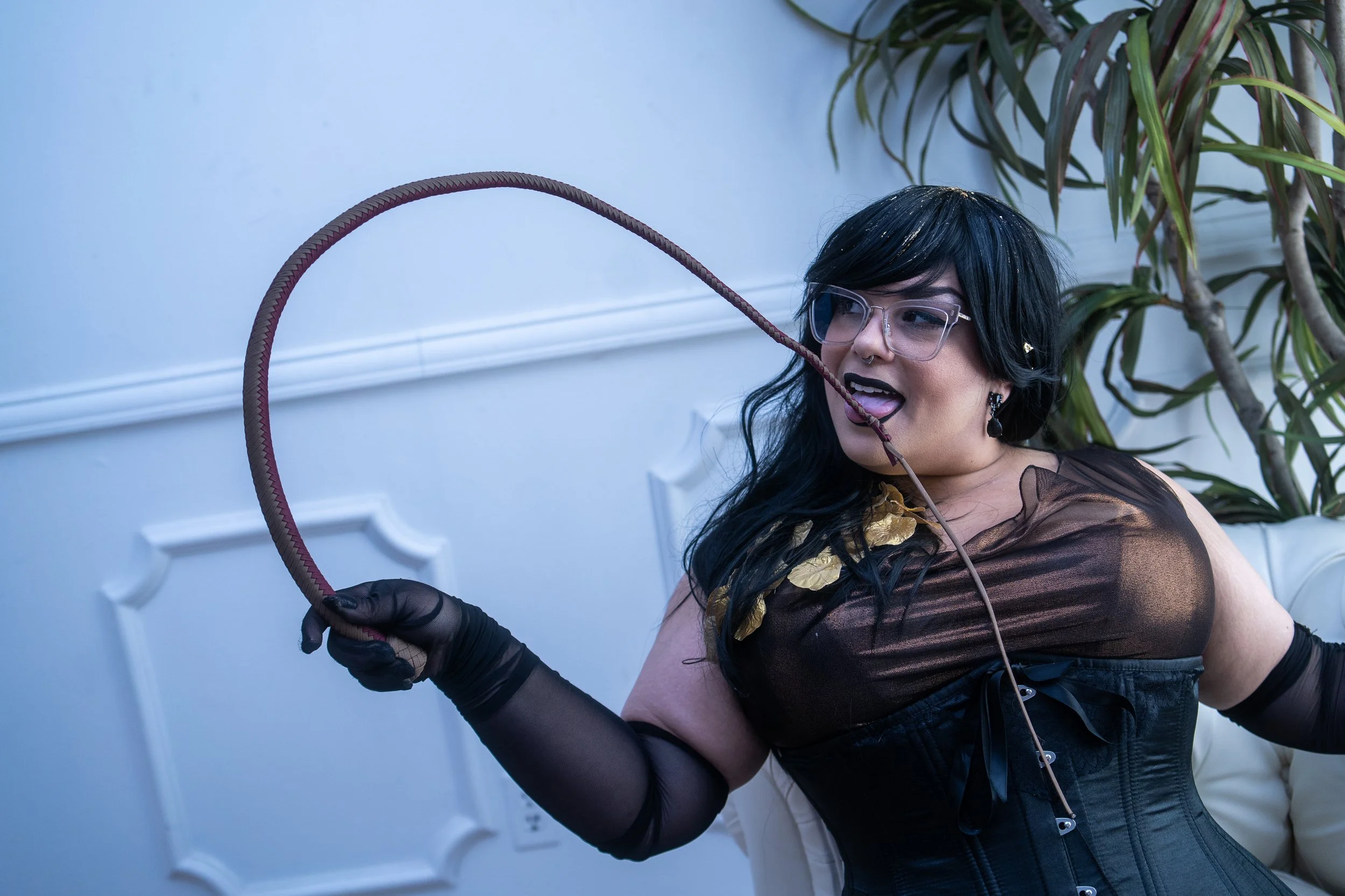 A woman with black hair, glasses, and dark makeup, wearing a black and gold dress, holding a whip, and demonstrating a playful or seductive pose indoors near a white wall and a tall plant.