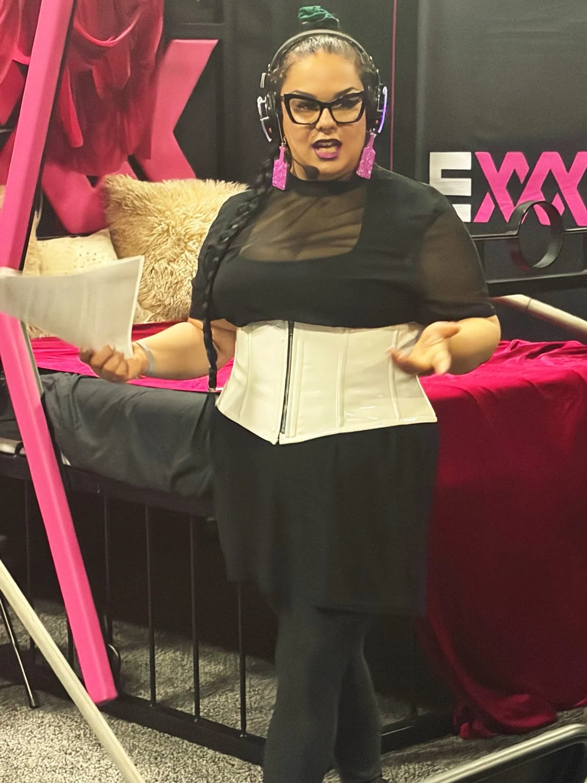 Pro Domme Dominatrix in Minneapolis with long braided hair, glasses, and purple earrings, wearing a black top with a see-through section and a white corset, giving a speech for a kink audience.