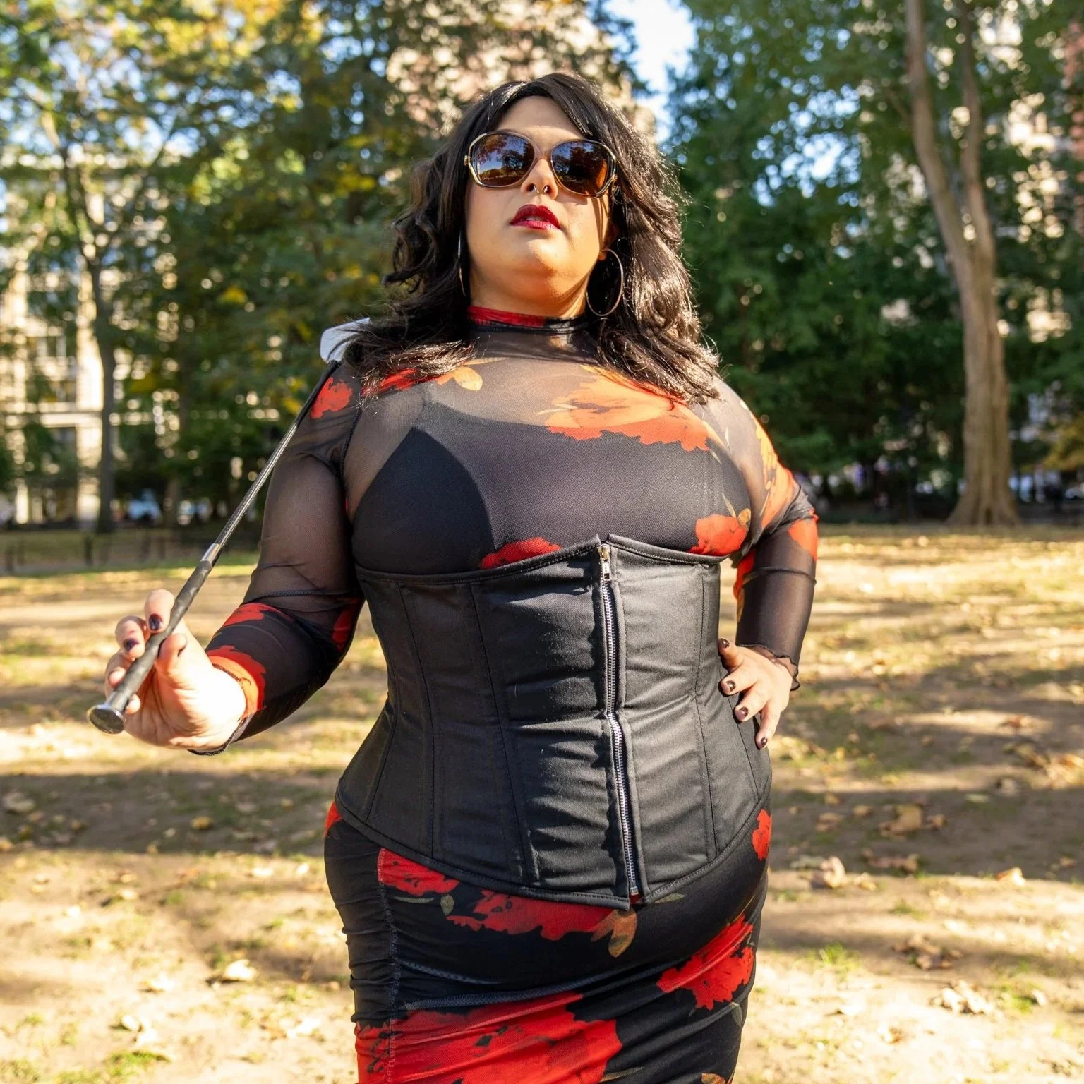 Madame Mona Pro Domme standing outdoors in a park, wearing sunglasses, a black leather corset, and a sheer black top. She has dark wavy hair, hoop earrings, and holding a selfie stick while looking at the camera. BDSM Dominatrix pro domme twin cities