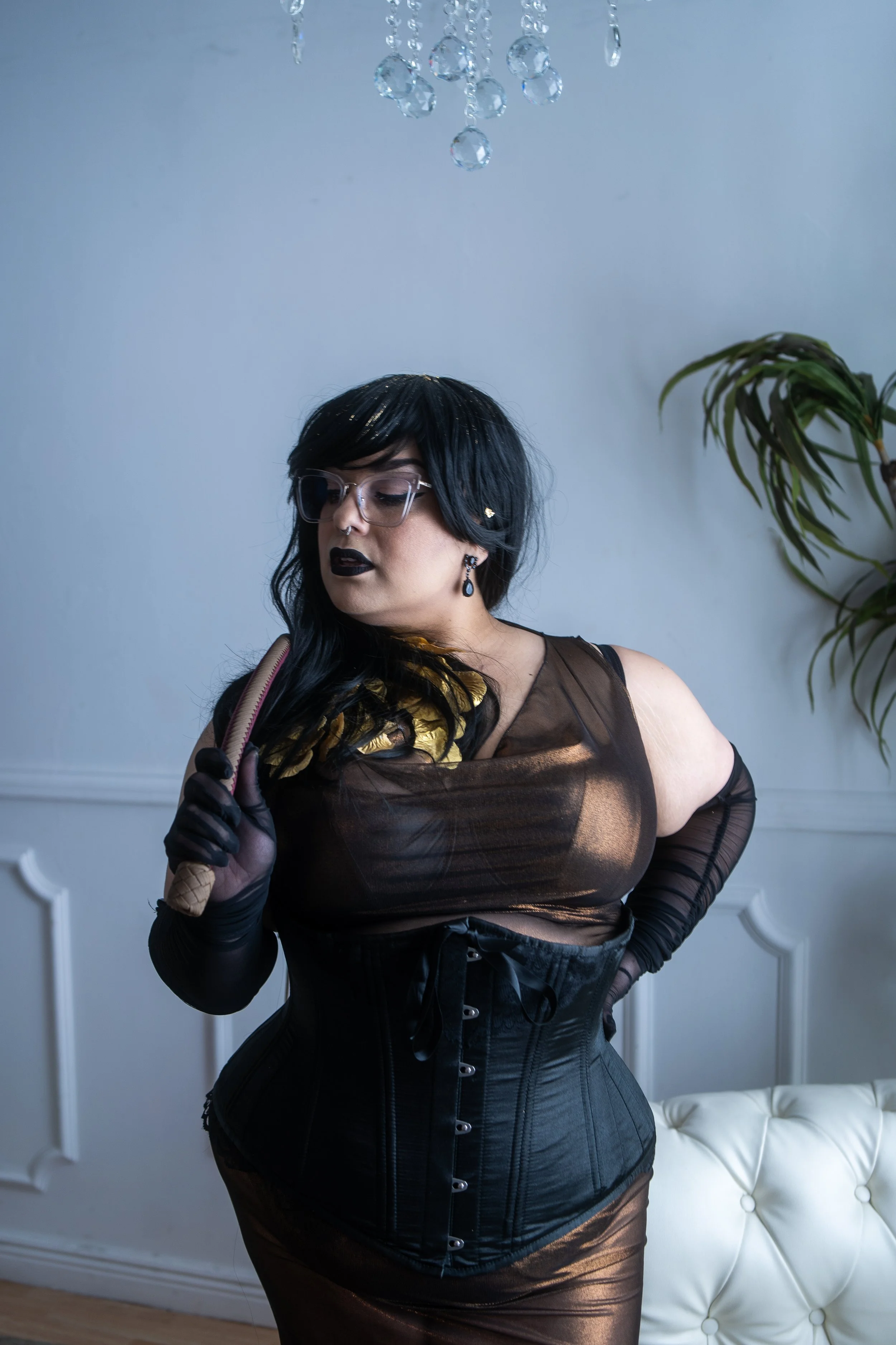 A woman with black hair, glasses, and dark lipstick wearing a sheer black and gold top and a black corset, holding an ice cream cone with a waffle cone, standing inside a room with white walls and a white tufted chair.