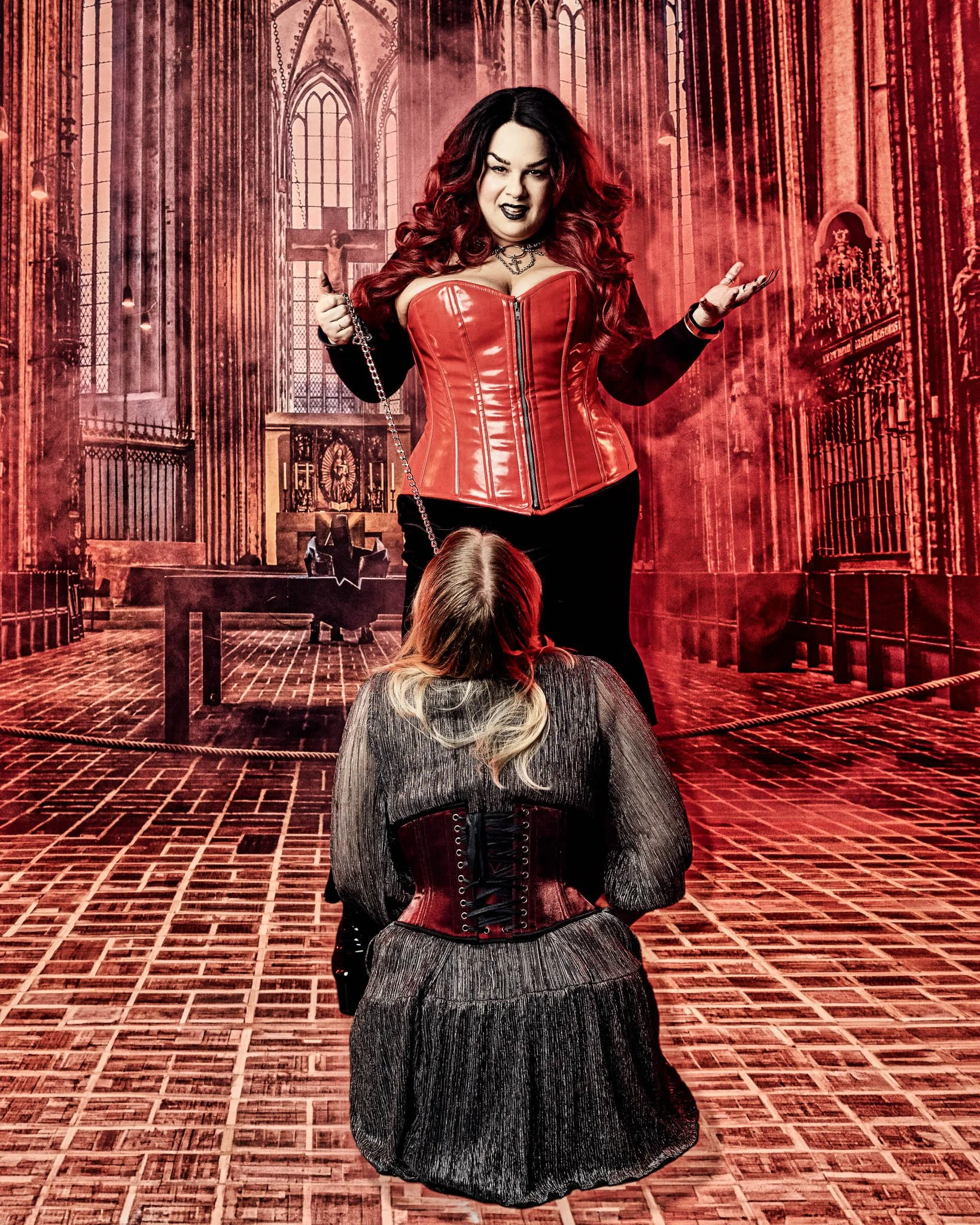 A woman dressed in gothic style with long dark hair and dark makeup, wearing a red latex corset and black lace-up skirt, stands in a church aisle with stained glass windows and religious statues, holding a chain with a crucifix.
