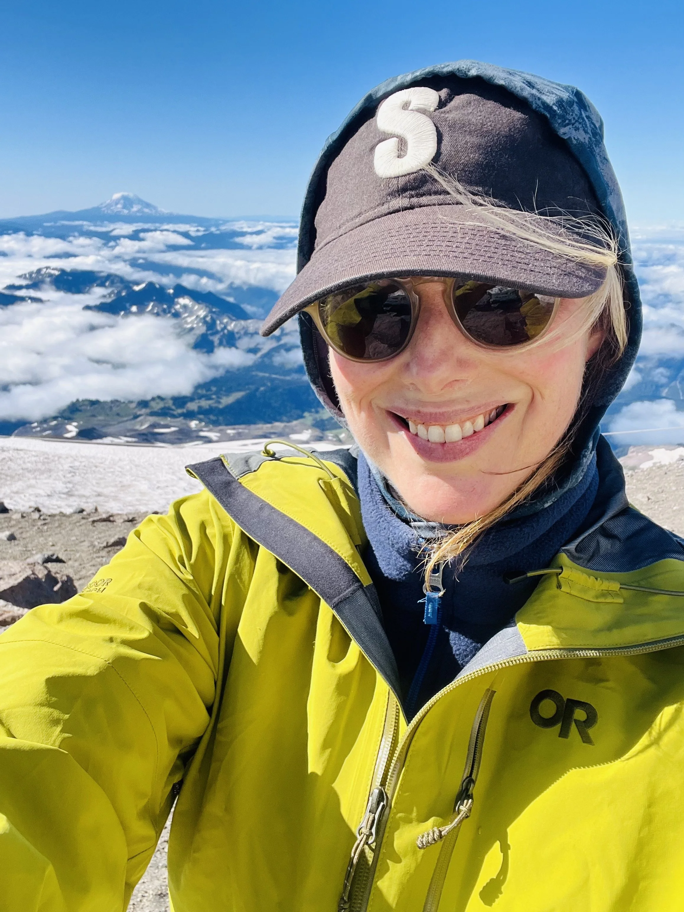 A person smiling outdoors on a snowy mountain with a snow-capped peak in the background, wearing a yellow jacket, sunglasses, and a cap.