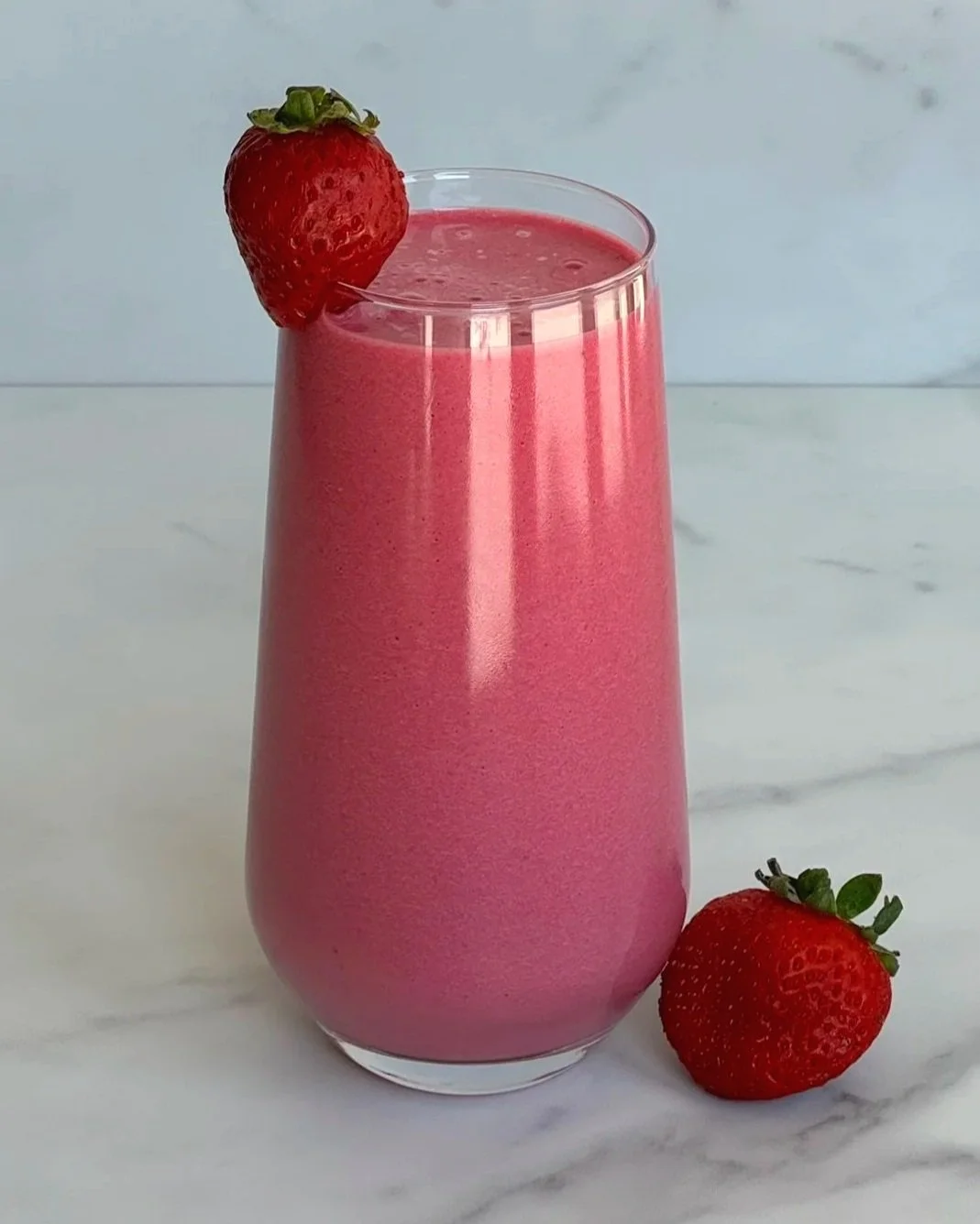 A tall glass of  Strawberry Banana Beet Protein Smoothie, garnished with a strawberry on the glass rim, and one strawberry at the base of the glass.