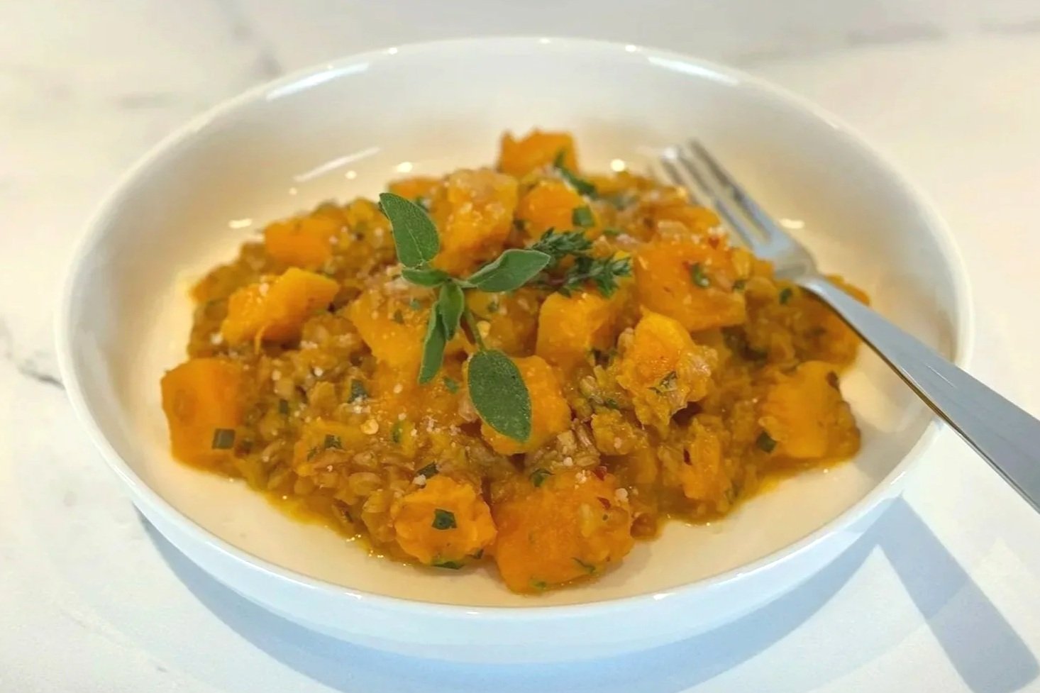 A white shallow bowl filled with Brown Butter Farro & Butternut Squash and topped with a touch of parmesan cheese & garnished with fresh sage and thyme.
