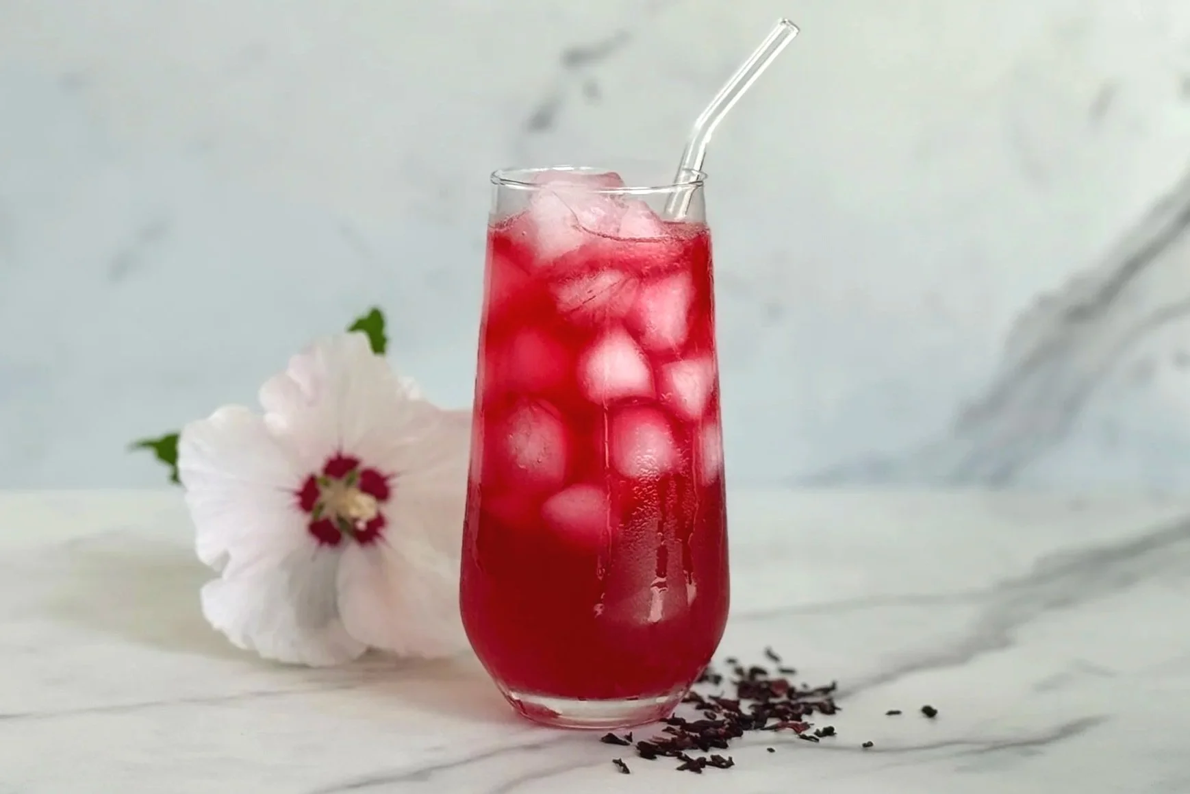 A tall highball glass of Hibiscus Lemonade with a glass straw. On the marble counter are a few dried hibiscus buds and 1 fresh hibiscus flower.