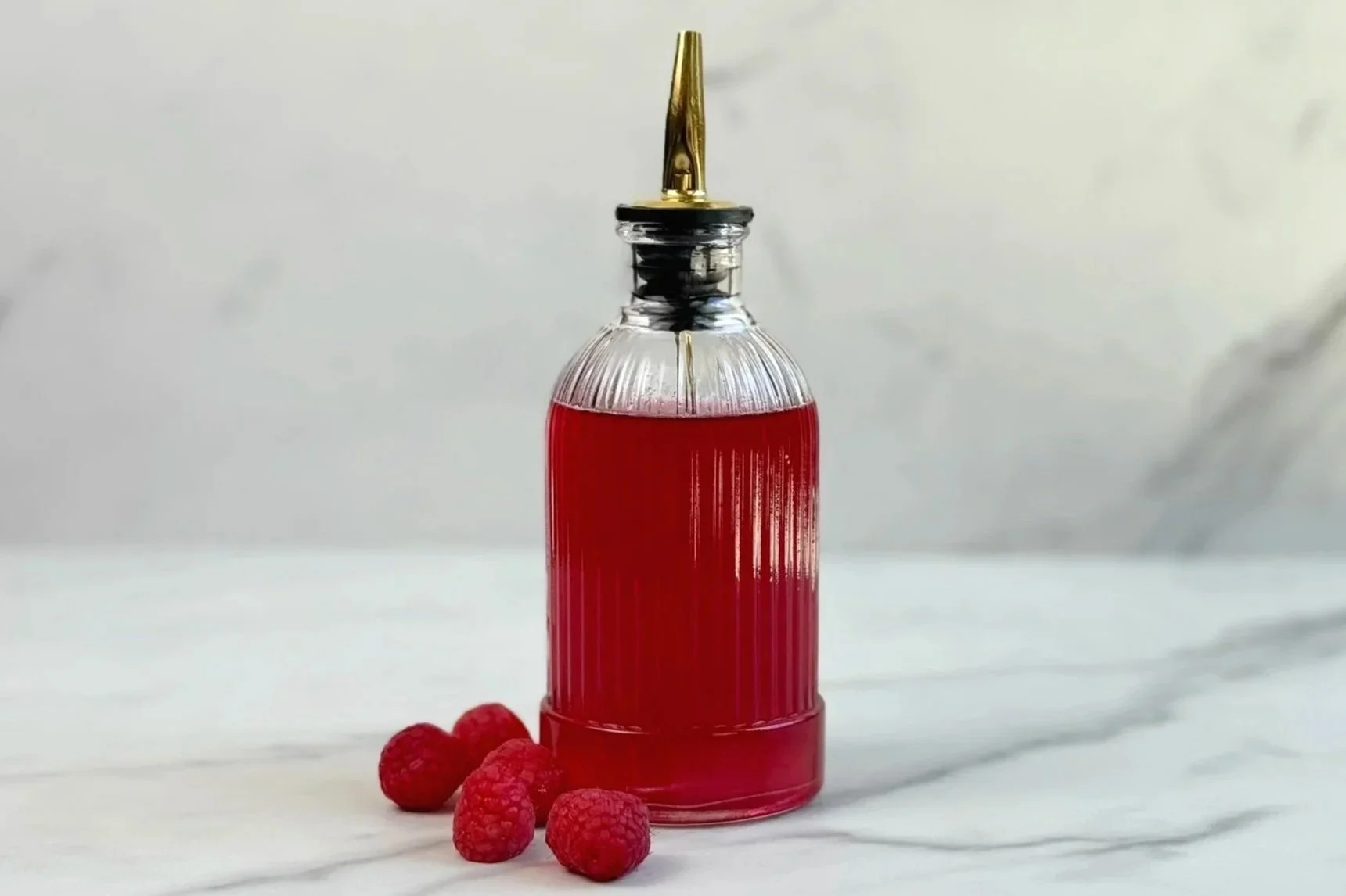 A bottle of Raspberry Simple Syrup with a pour spout is sitting on a marble countertop, surrounded by fresh raspberries.