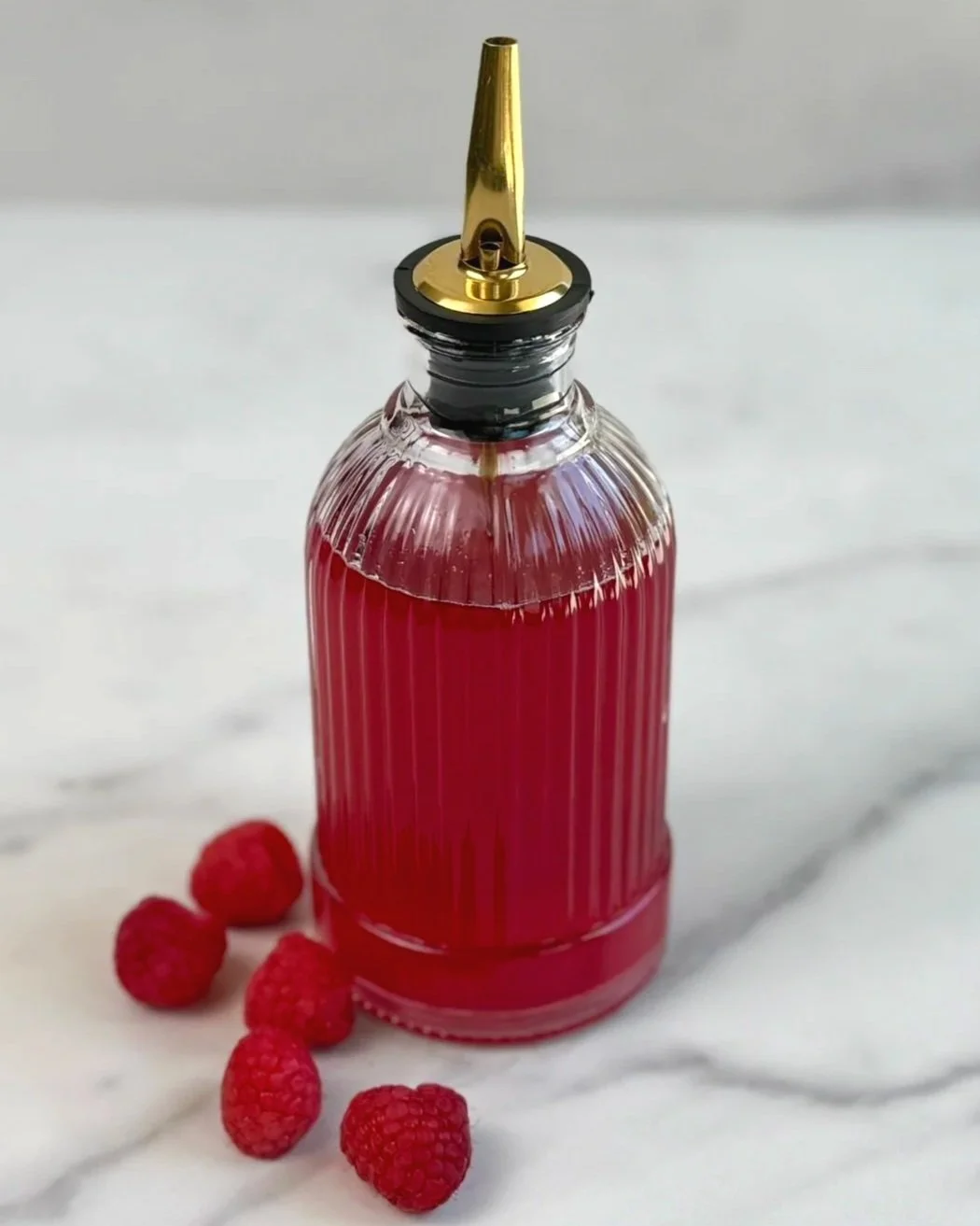 A bottle of Raspberry Simple Syrup with a pour spout is sitting on a marble countertop, surrounded by fresh raspberries.