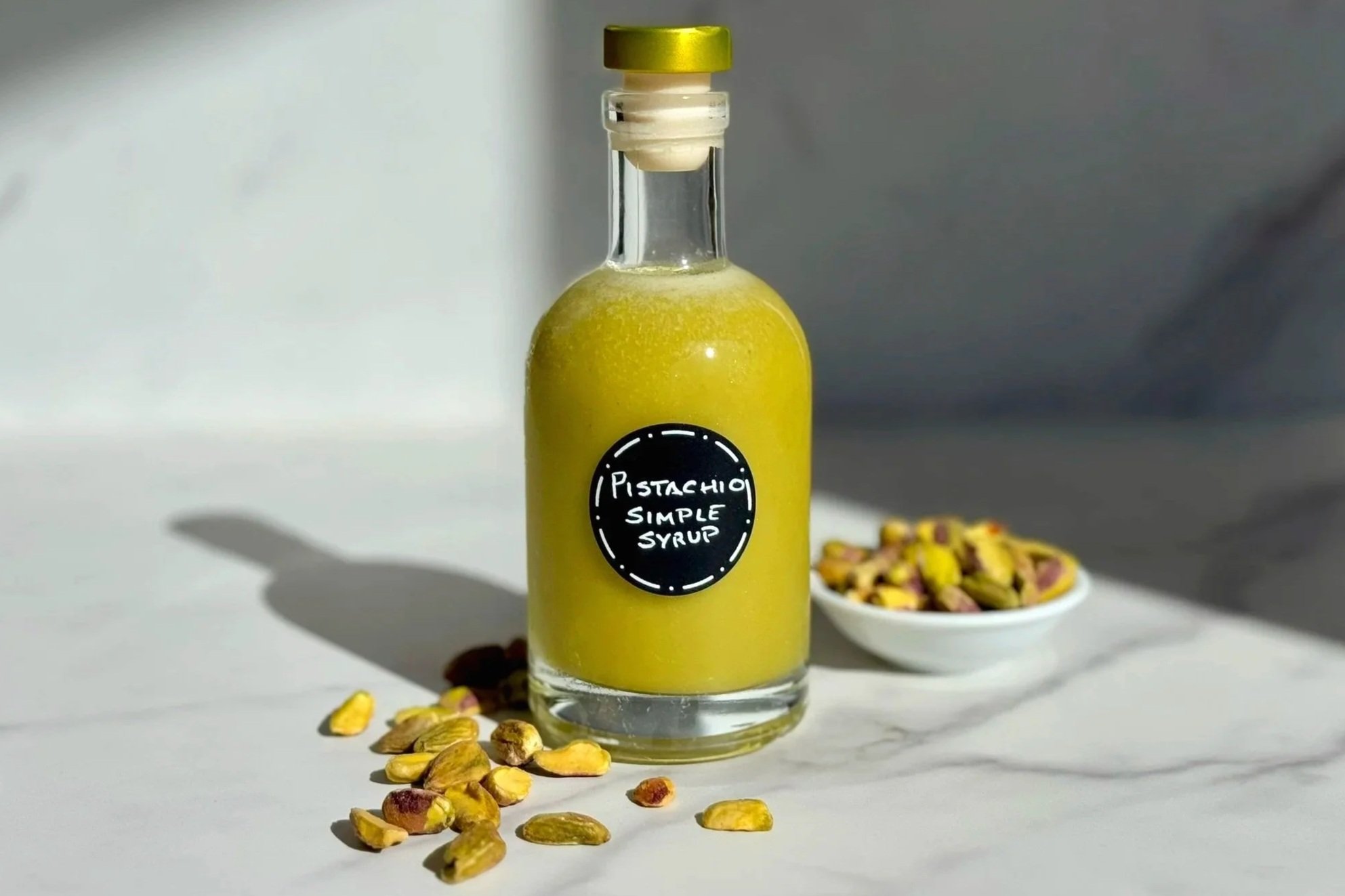 A glass bottle of Robust Pistachio Simple Syrup is sitting on a marble counter with a handful of shelled pistachios scattered at the base of the bottle. A small dish of pistachio nuts sit in the background.