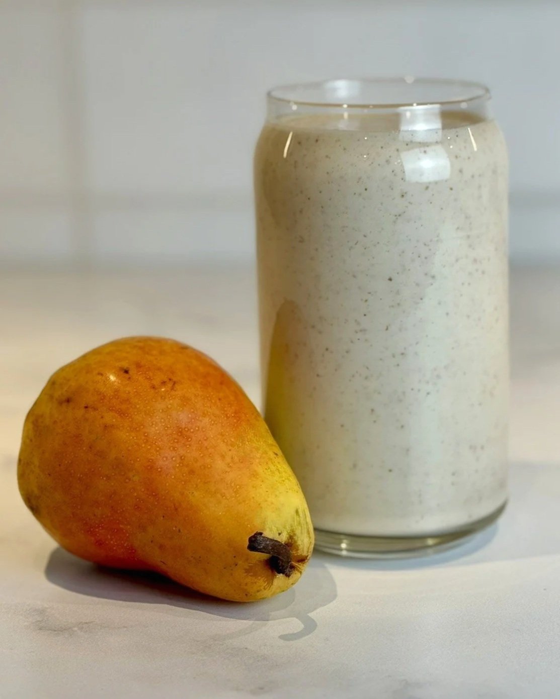 A glass of High Protein Pear Ginger Smoothie sitting on a counter next to a fresh Bartlett pear.