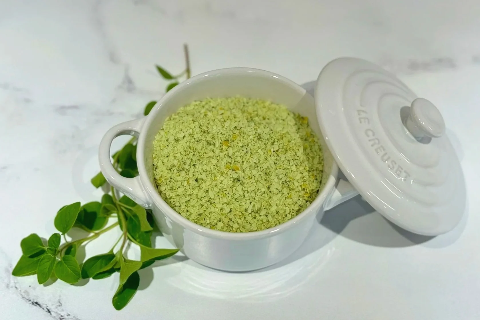 A white Le Creuset salt keeper filled with Lemon Oregano Salt. A whole lemon and a sprig of fresh oregano are resting on the counter next to it.