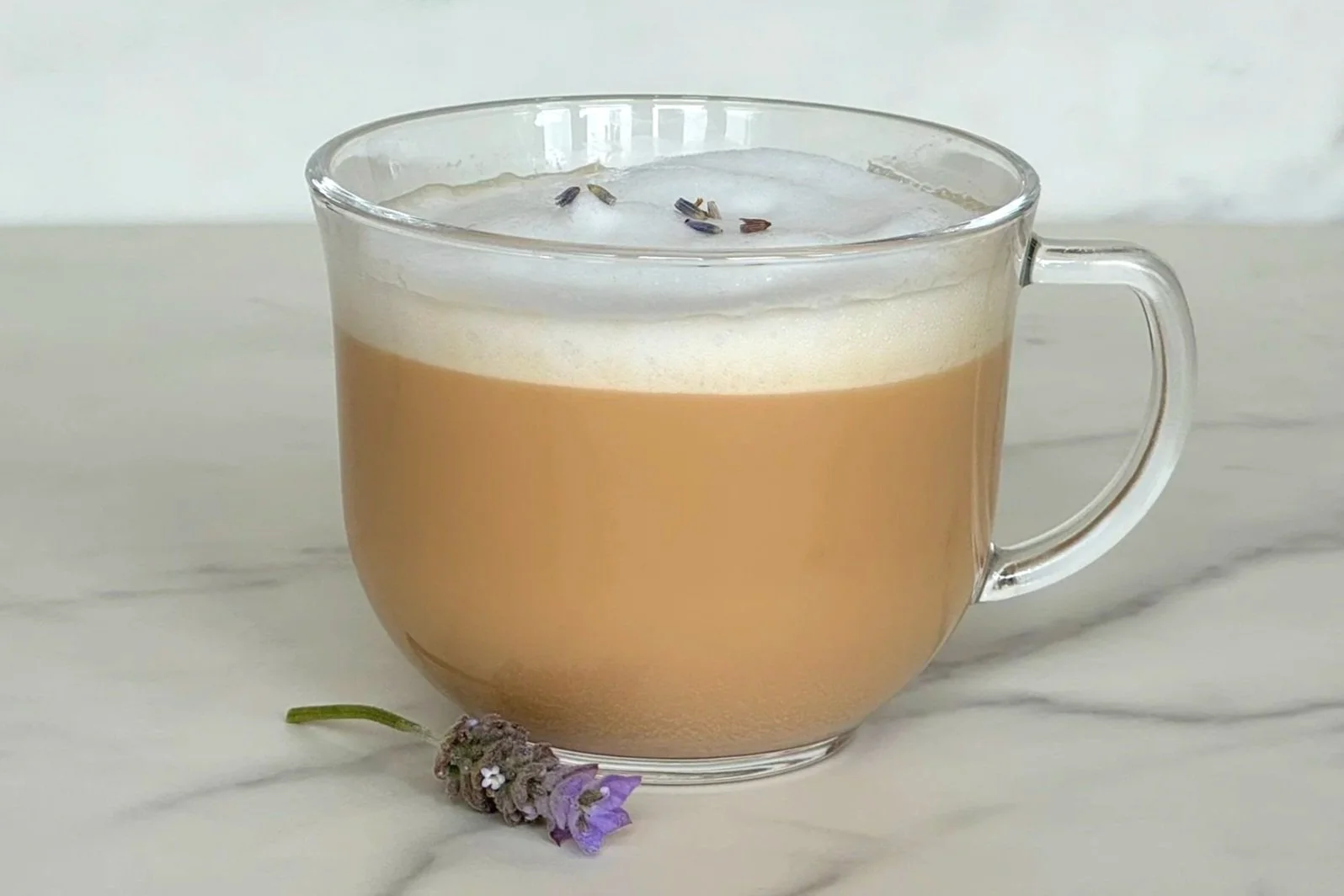 A clear glass mug of rich and foamy Lavender London Fog sitting on a counter with a single lavender flower bud resting on the counter.