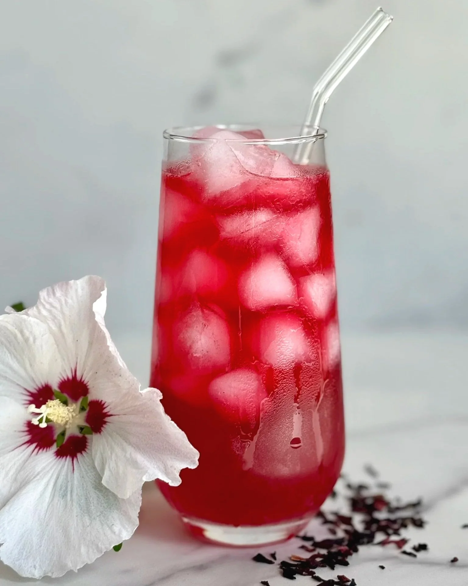 A tall highball glass of Hibiscus Lemonade with a glass straw. On the marble counter are a few dried hibiscus buds and 1 fresh hibiscus flower.