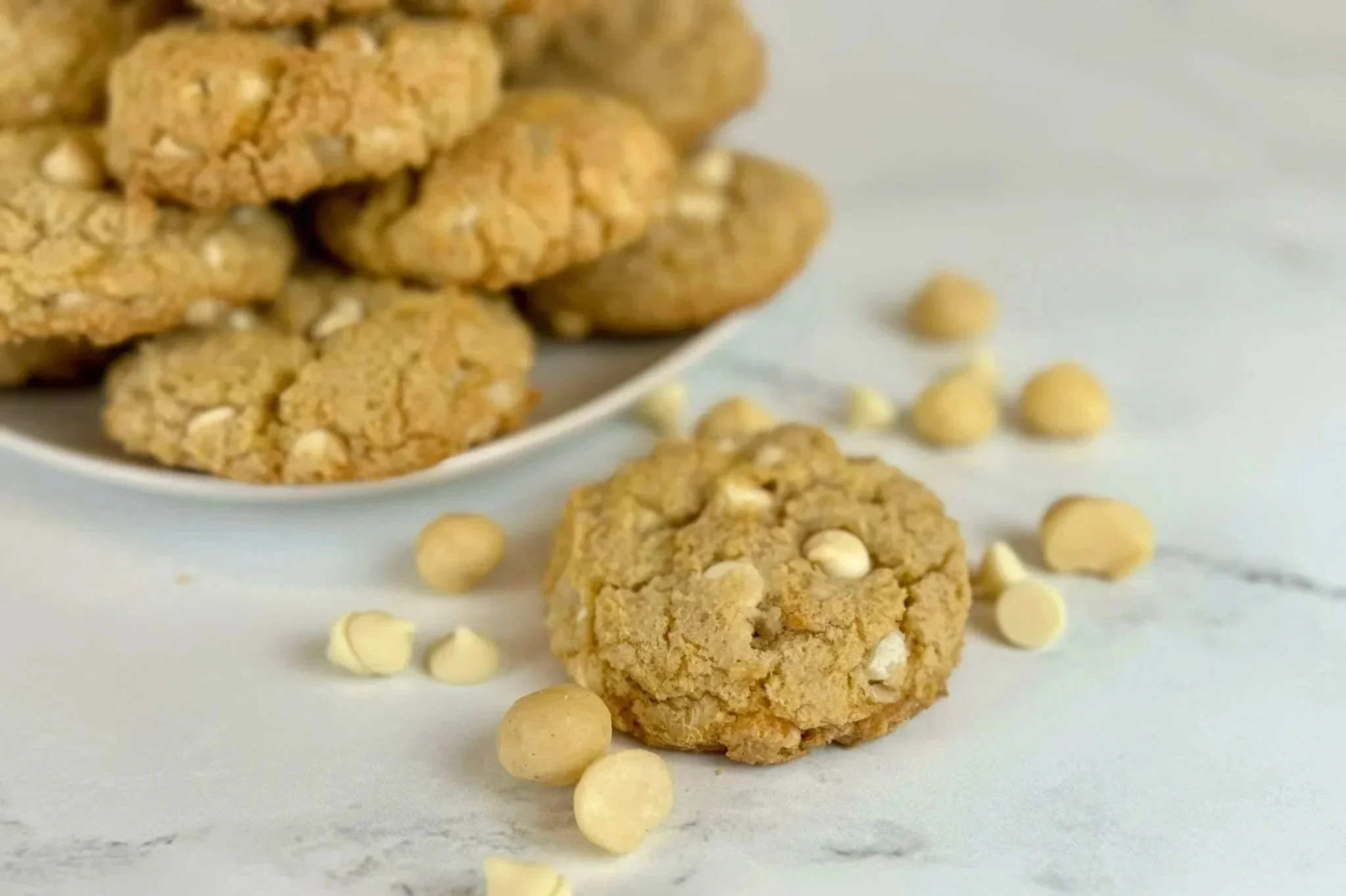A plate piled high with Chewy White Chocolate Macadamia Nut Cookies. One cookie sits on the counter with a few sprinkles of white chocolate chips and macadamia nuts resting on the counter.