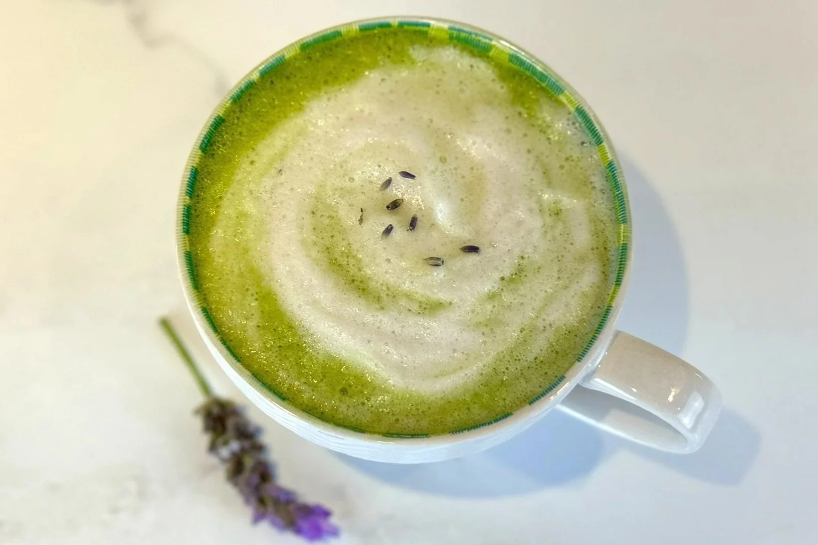 A large rounded white mug filled with a foamy Lovely Lavender Matcha Latte that is garnished with lavender buds. A lavender flower is resting on the counter next to the mug.