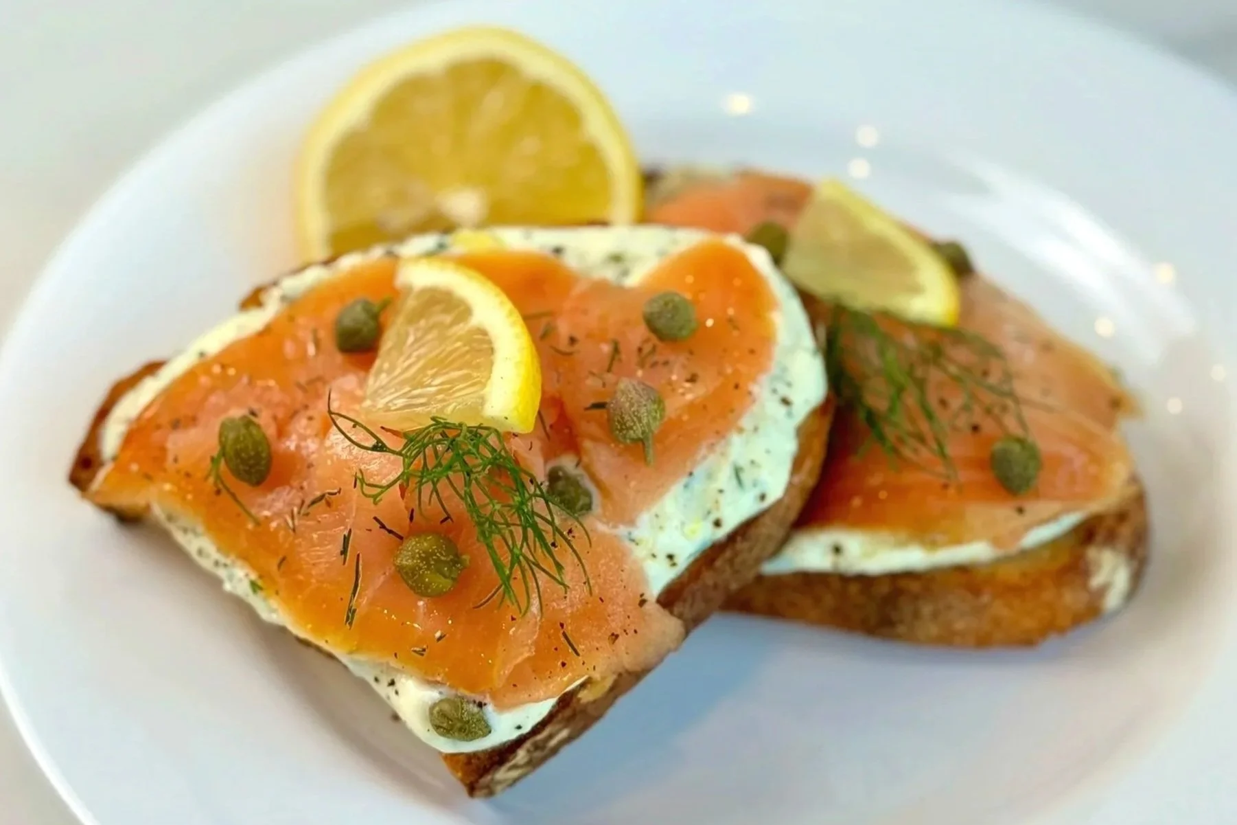 A white plate with two pieces of Smoked Salmon Toast with Lemon Whipped Ricotta that are garnished with capers, sprig of dill, and thinly sliced lemon.