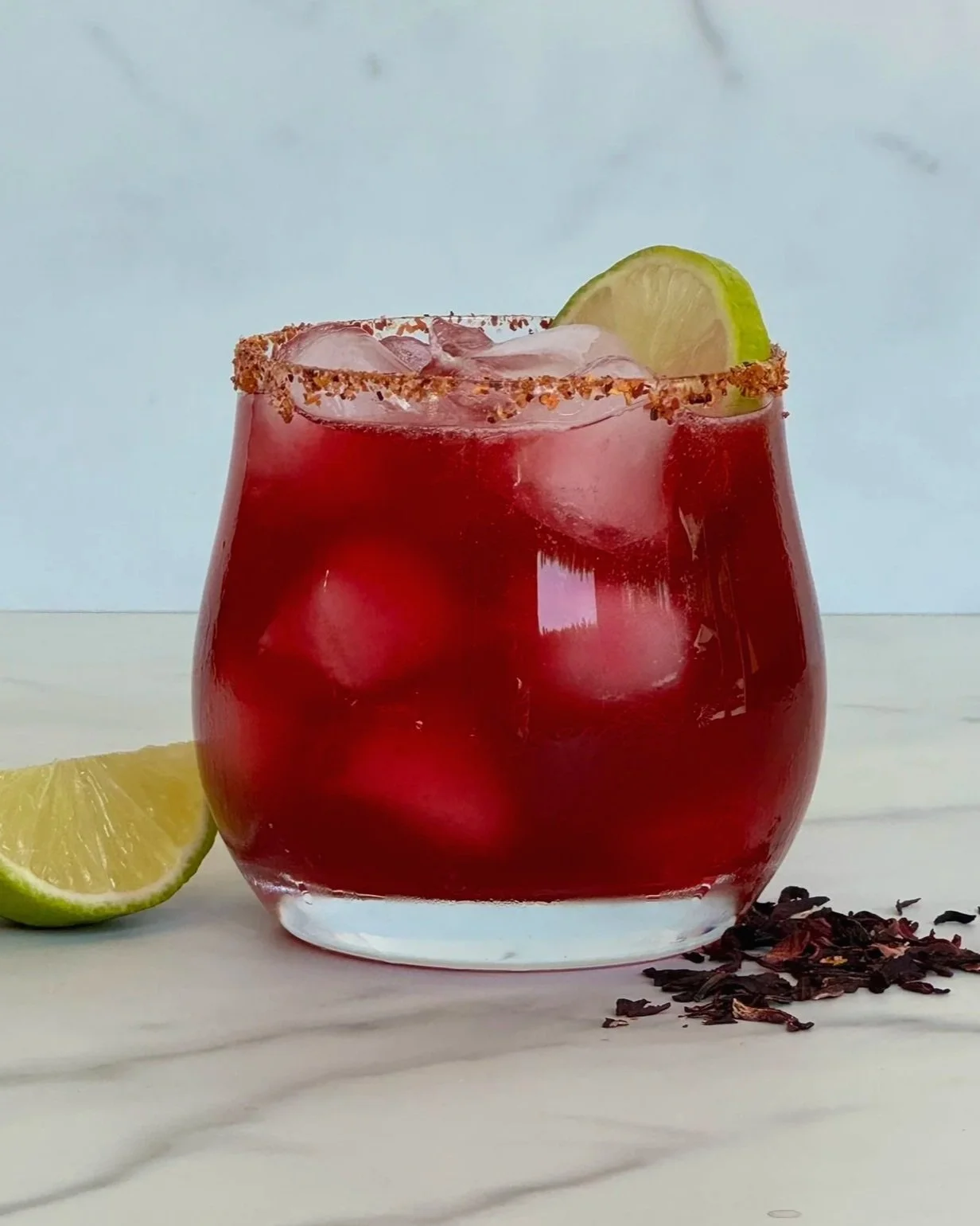 A cold cocktail glass of Hibiscus Mezcal Margarita that is garnished with a Tajin rim and lime slice. A wedge of lime and some dried hibiscus buds are scattered on the counter.