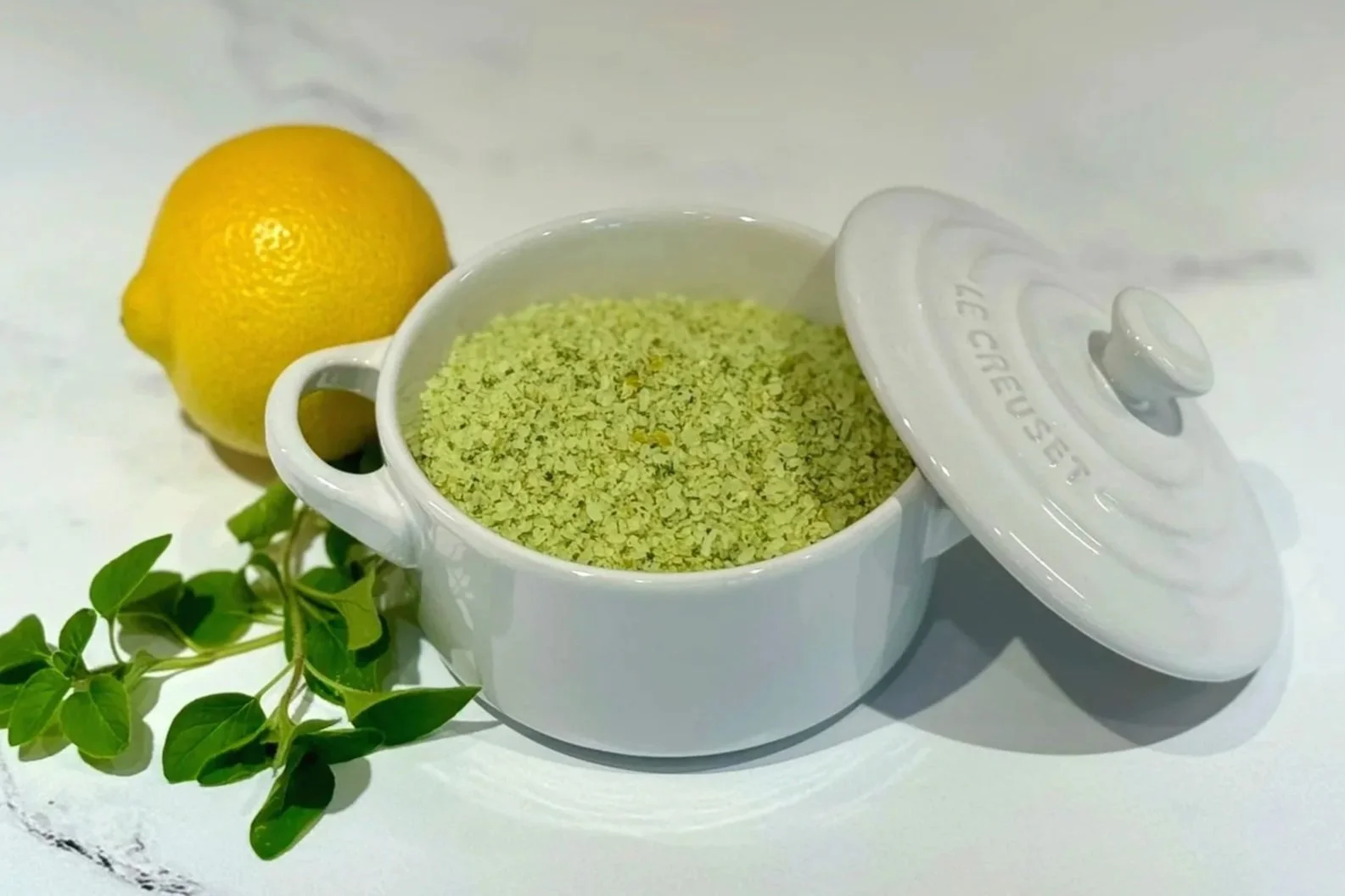 A white Le Creuset salt keep filled with Lemon Oregano Salt. A whole lemon and a sprig of fresh oregano are resting on the counter next to it.